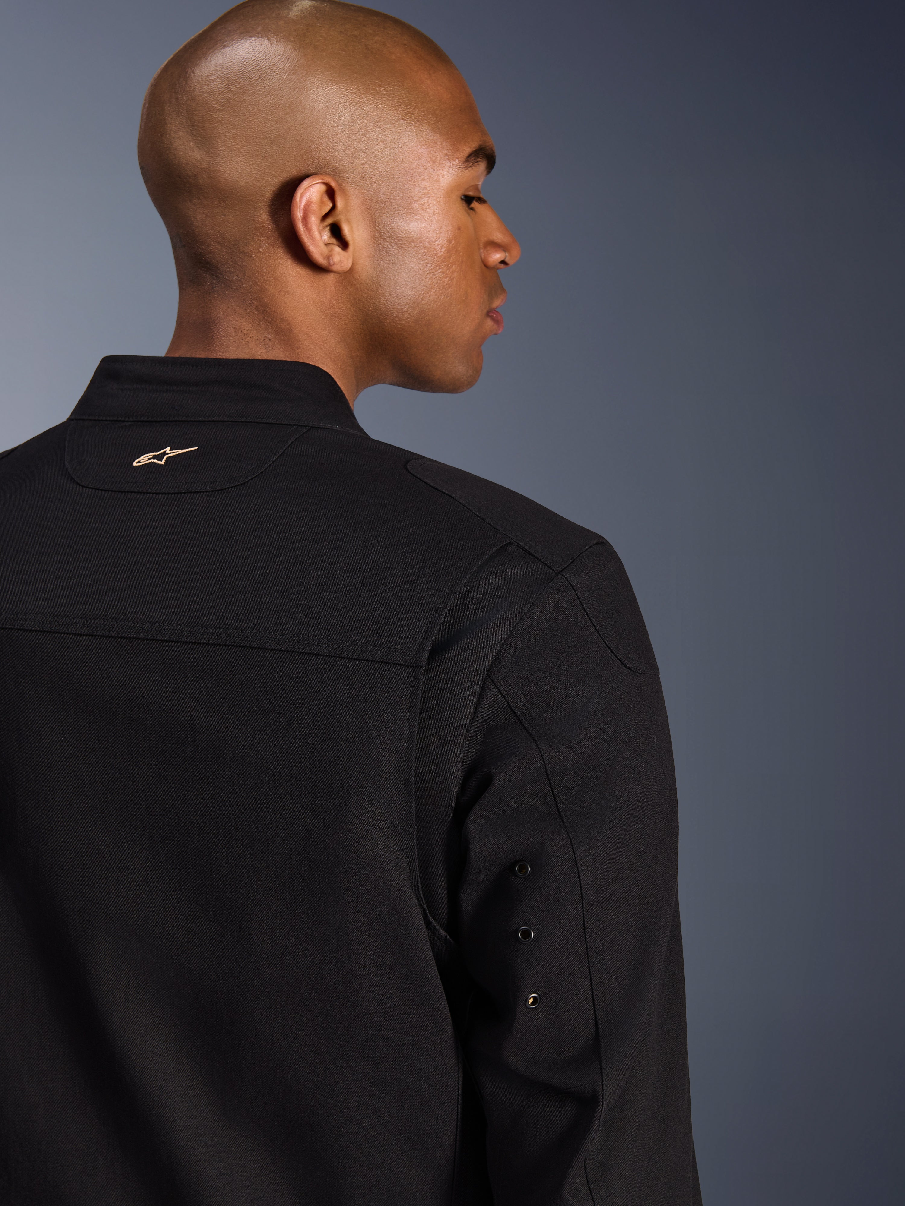 Alpinestars Bruiser Jacket, textile urban motorcycle jacket, Black, close-up back view featuring an embroidered gold logo on the upper back, metal eyelet sleeve details, and pleated shoulder panels for enhanced mobility.