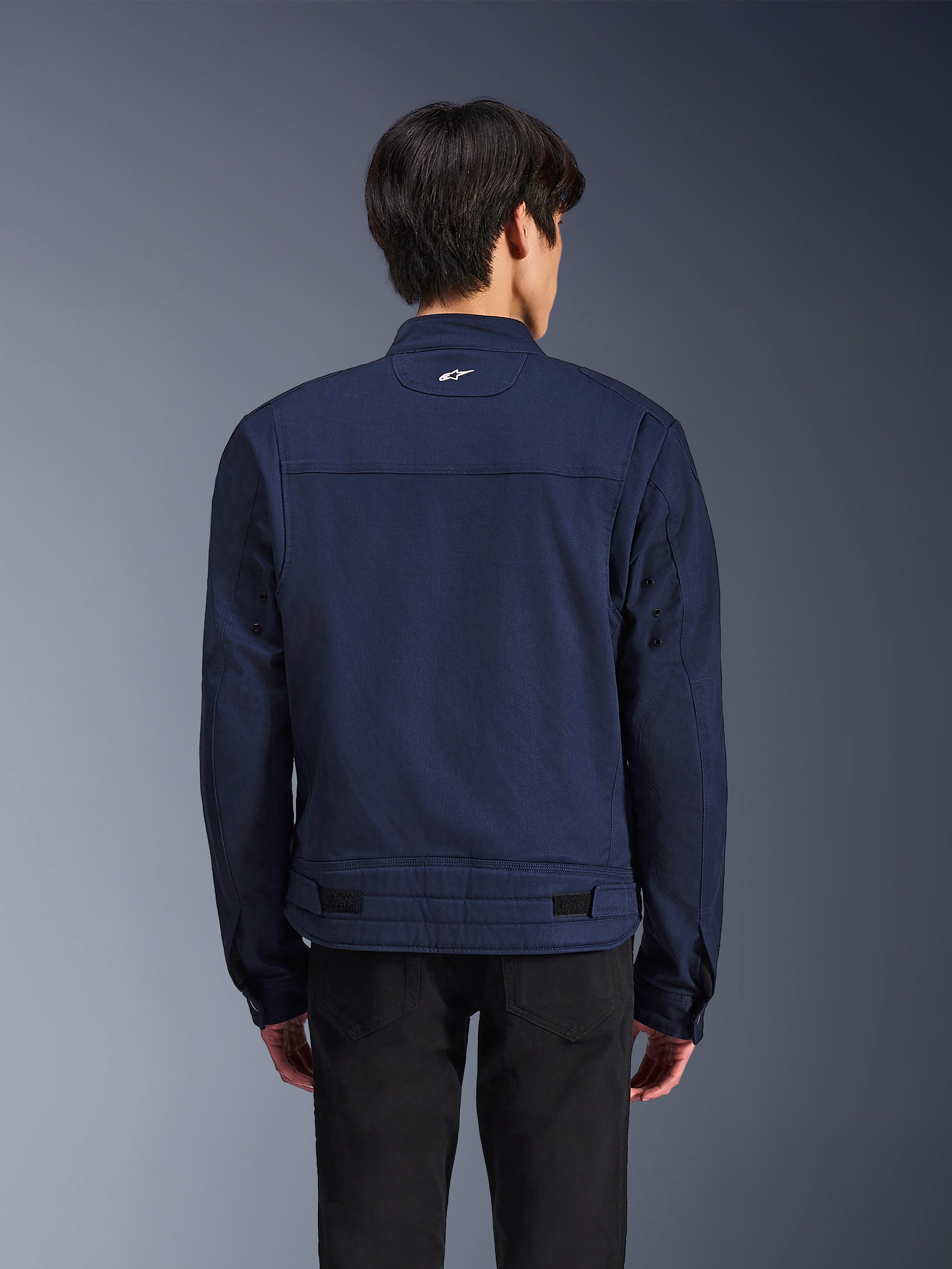 Alpinestars Bruiser Jacket, Dark Blue textile motorcycle jacket, back view on model, workwear-inspired design with a small white Astars logo on the neck, adjustable waist tabs, urban contemporary riding apparel.