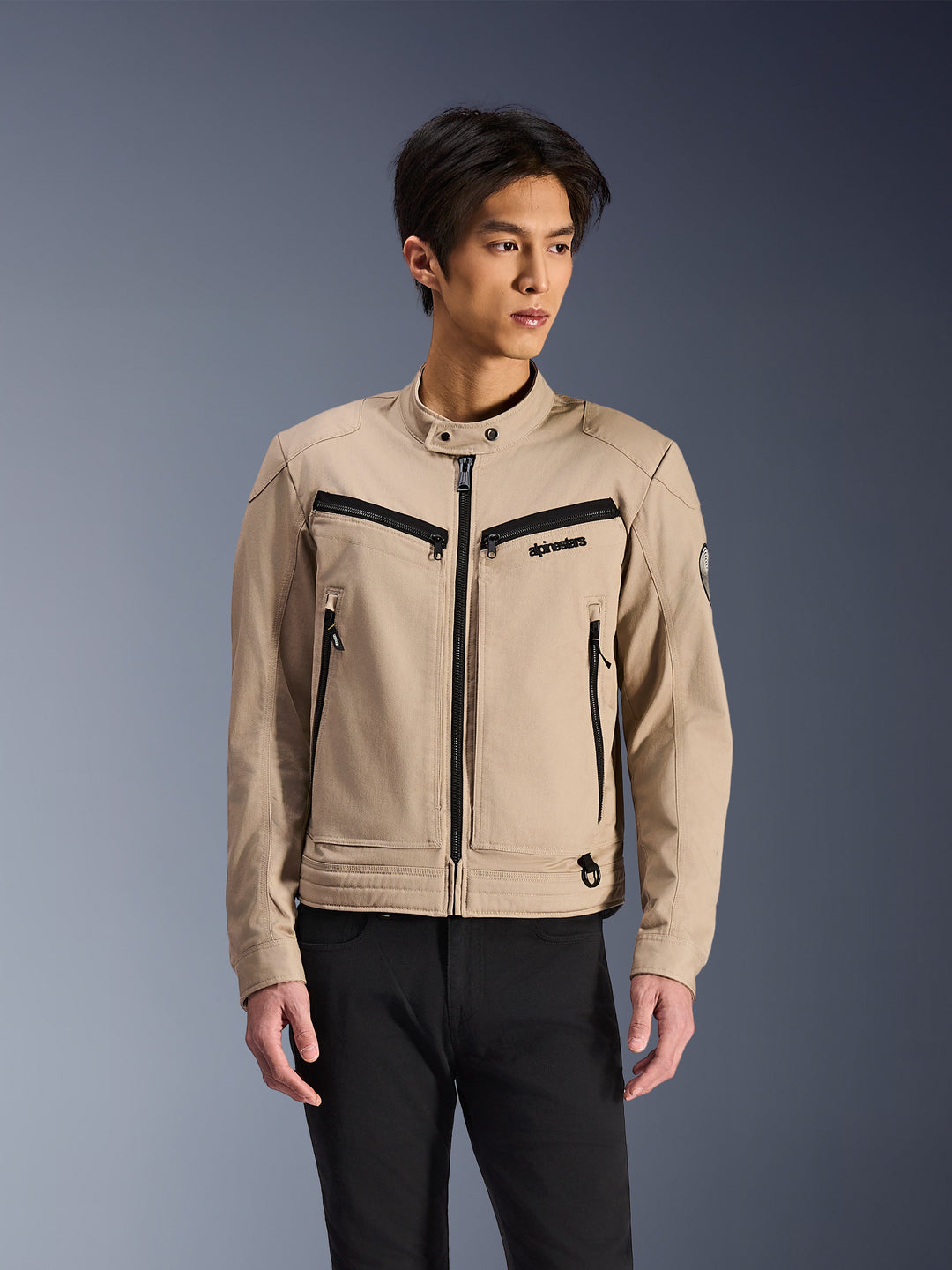 Alpinestars Bruiser Jacket, Beige motorcycle jacket worn by male model, featuring black zipper details on chest and side pockets, high snap collar, workwear-inspired urban riding wear, paired with black pants
