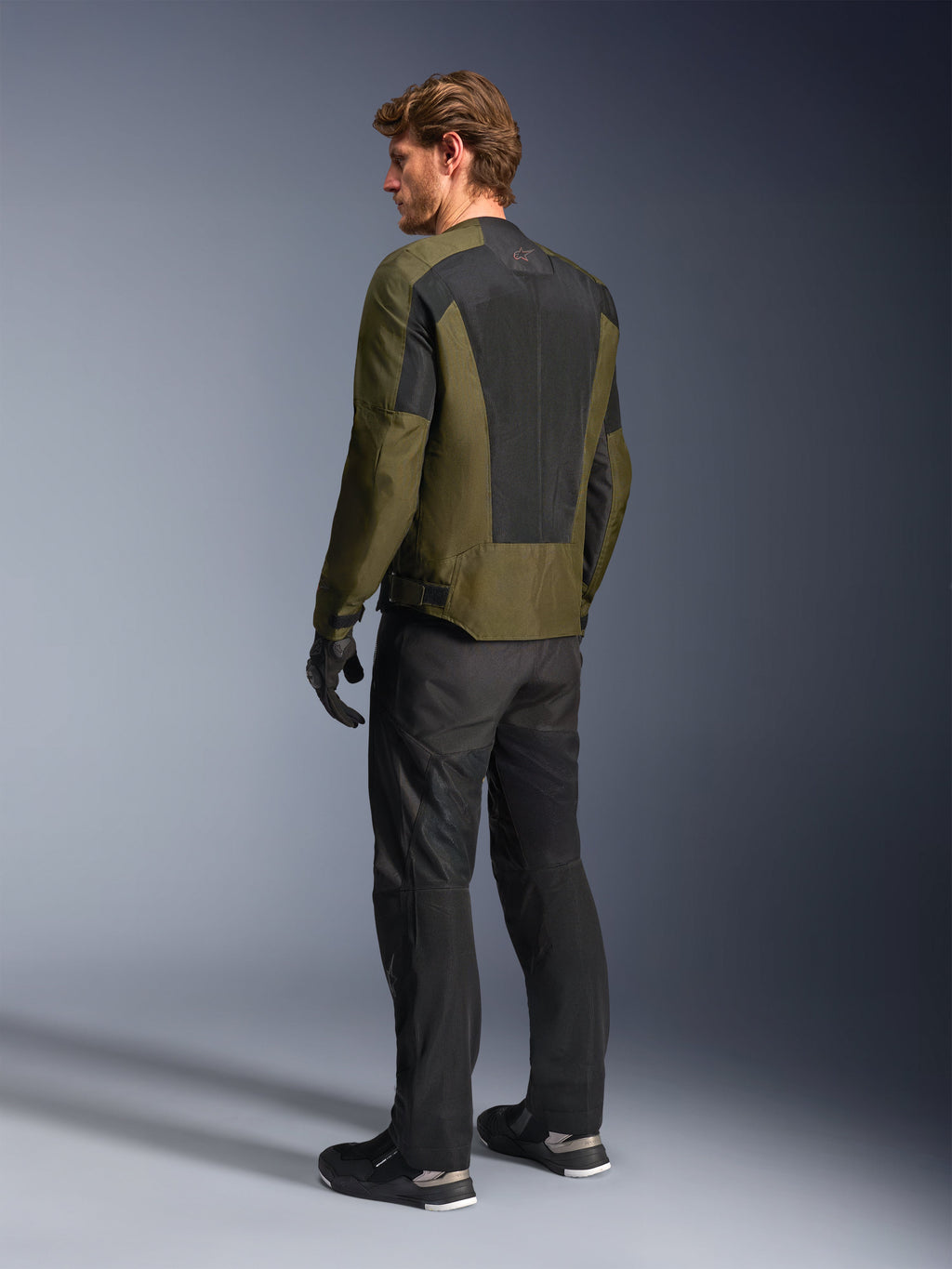Male model wearing Alpinestars C-1 Air Jacket in Khaki, urban motorcycle jacket, rear view highlighting large black mesh panels on the back, paired with black technical riding pants and gloves, contemporary motorcycle gear for warm weather
