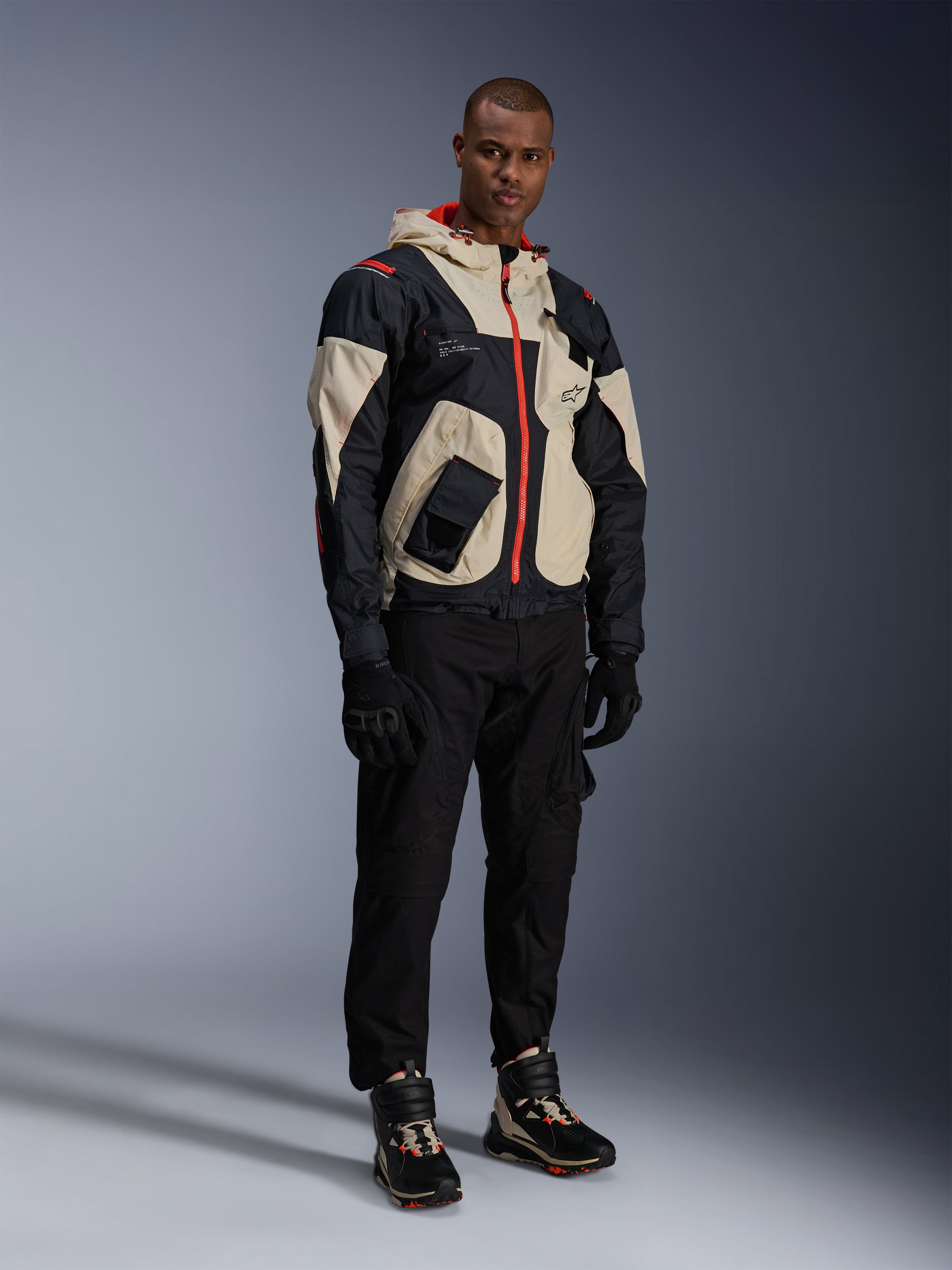 Model wearing the Alpinestars Mogress Air Jacket in Black, Almond, and Hot Orange, urban motorcycle jacket with a utility-inspired design, worn with black riding pants, black gloves, and coordinated technical boots, contemporary street-ready riding gear