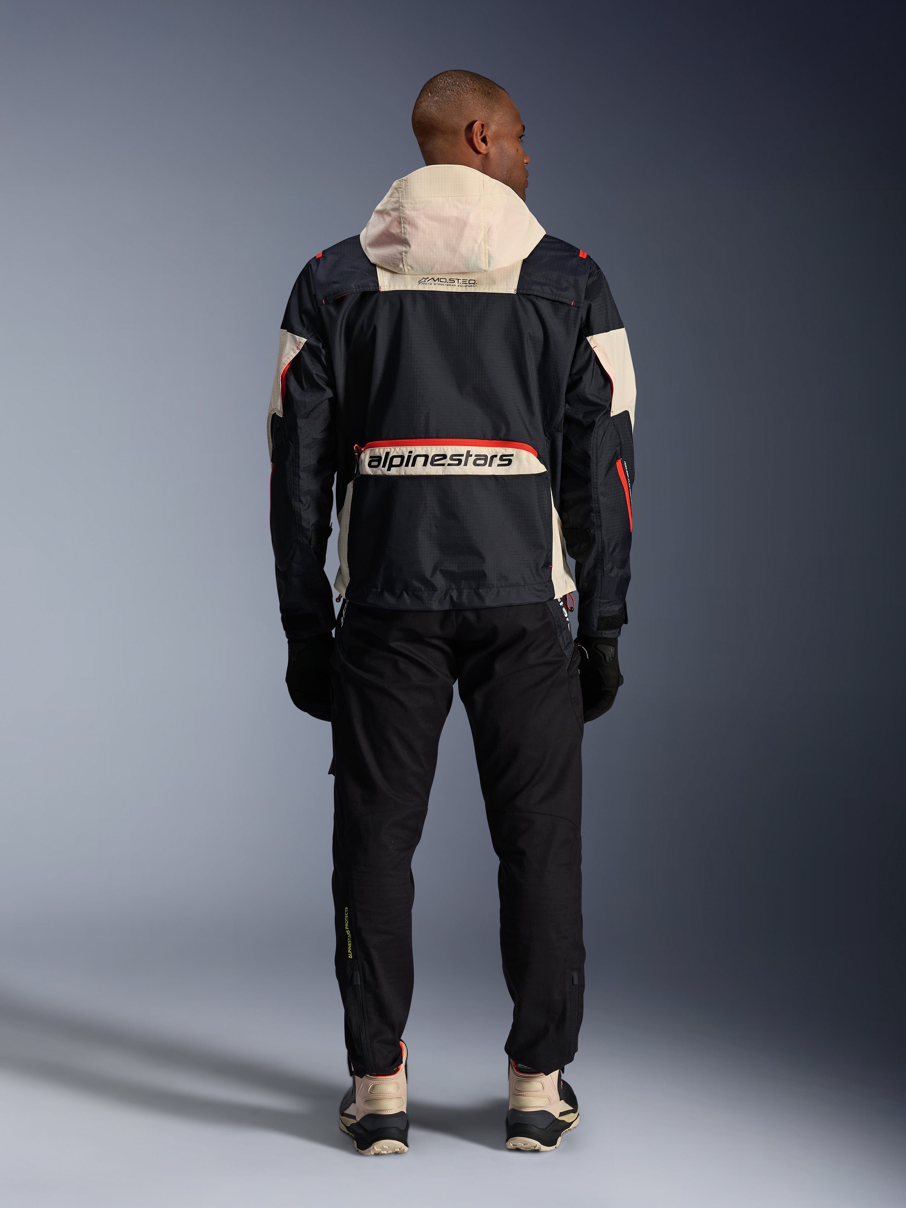 Rear view of a rider wearing the Alpinestars Mogress Air Jacket in Black, Almond, and Hot Orange, featuring a cream-colored hood, large Alpinestars branding across the back pocket, and orange zipper accents, paired with black pants and cream boots for an urban motorcycle look.