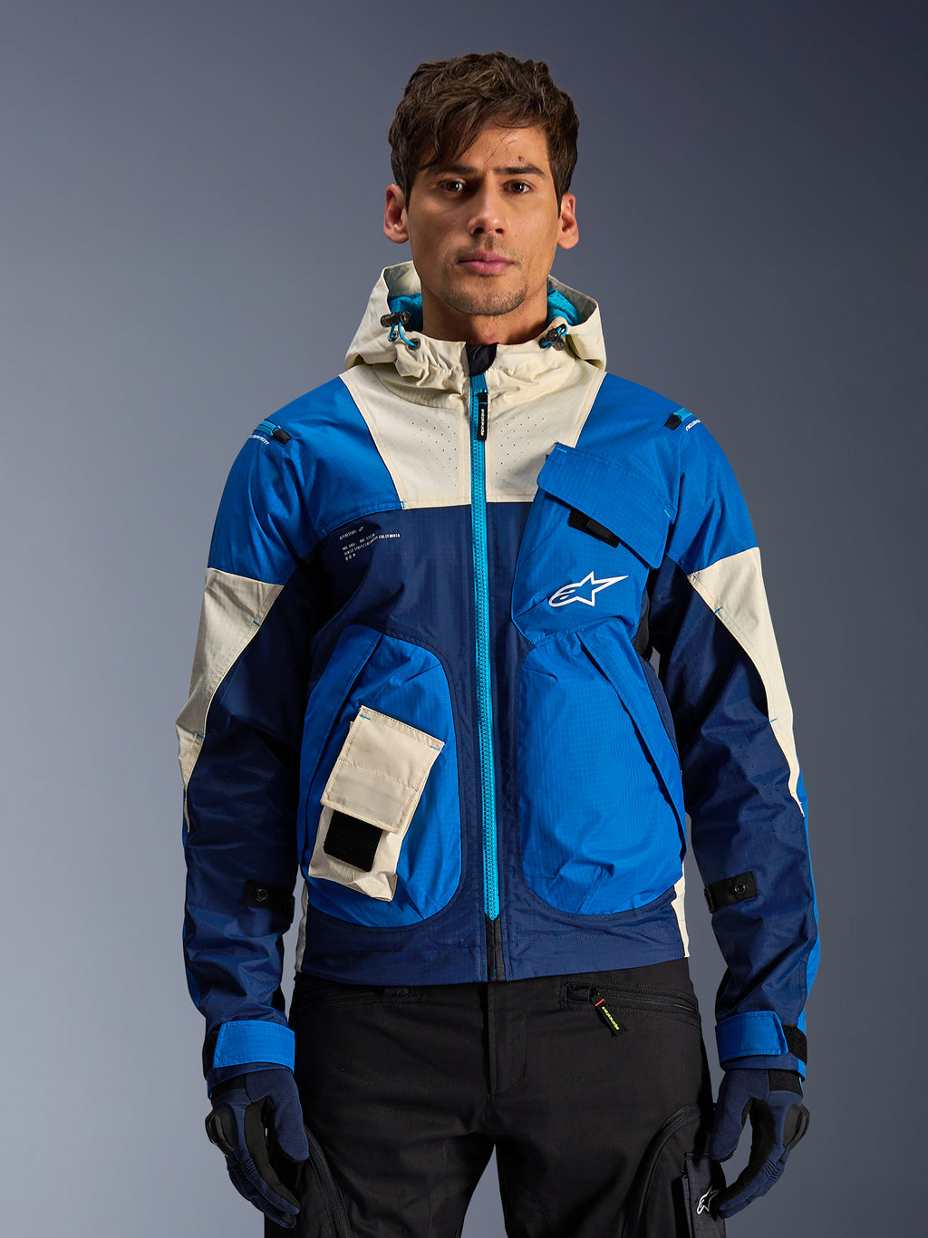 Alpinestars Mogress Air Jacket in Blue Almond, hooded urban motorcycle jacket with contrasting blue and off-white panels, utility pockets and teal front zipper, worn by a model with black pants and gloves