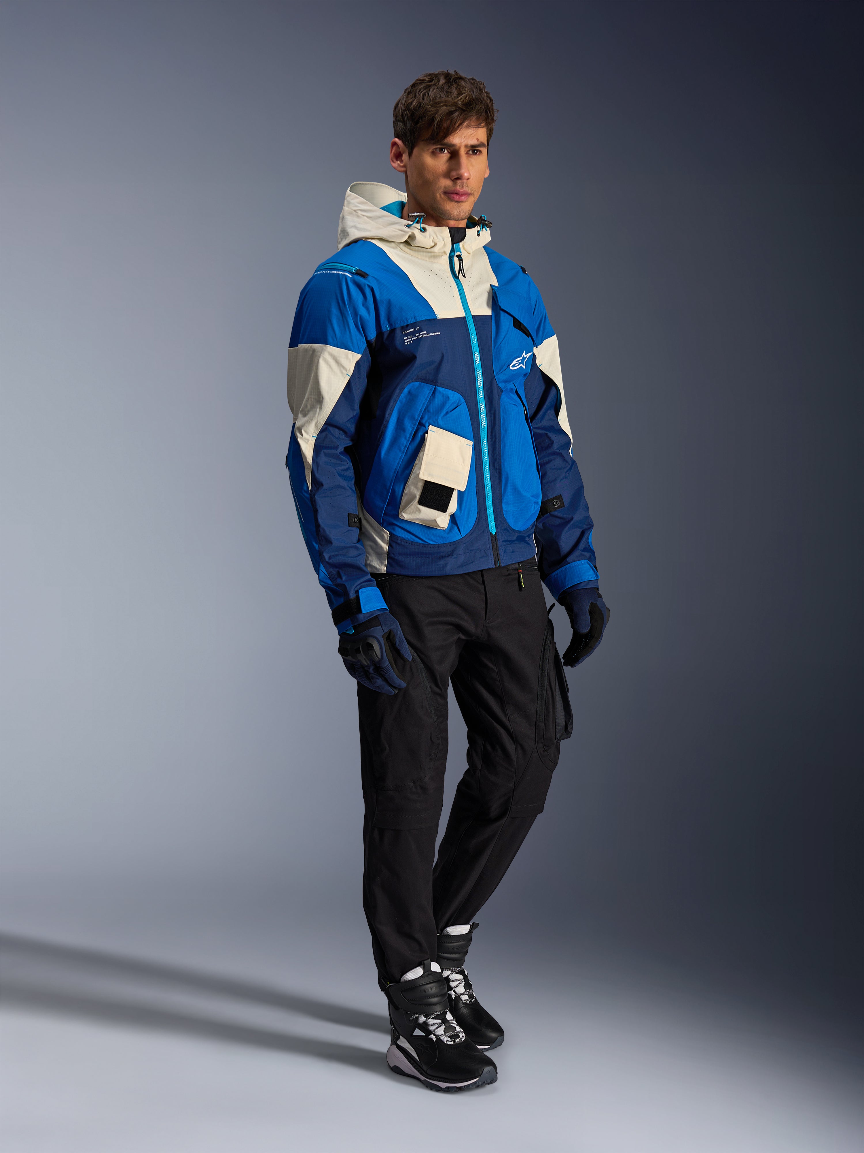 Alpinestars Mogress Air Jacket, Urban Motorcycle Jacket, Blue Almond with cream and navy panels, featuring a hood and utility pockets, worn with black riding pants, gloves, and boots, contemporary streetwear-inspired protective gear.