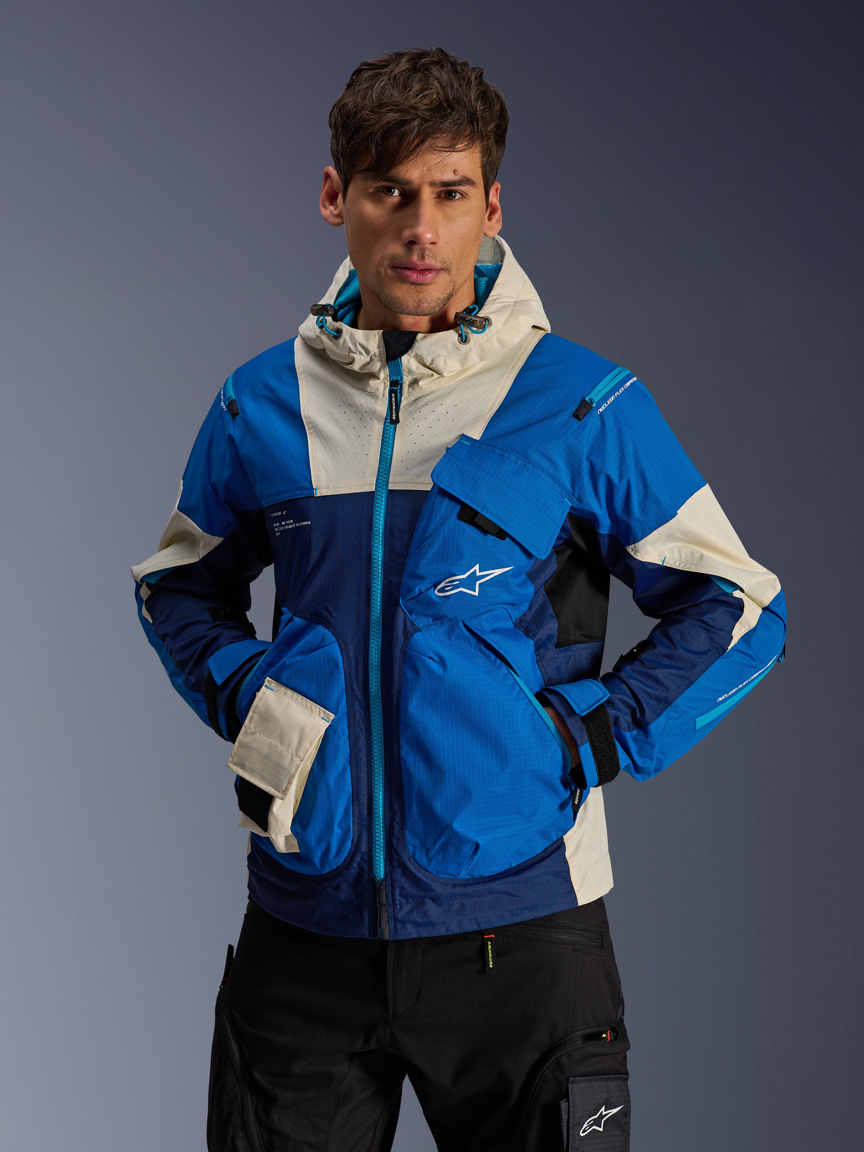 Male model wearing the Alpinestars Mogress Air Jacket in Blue Almond, urban motorcycle jacket with bright blue and navy color-blocking, cream accents on the hood and pockets, technical ripstop fabric with utility pockets, modern motorsport-inspired streetwear
