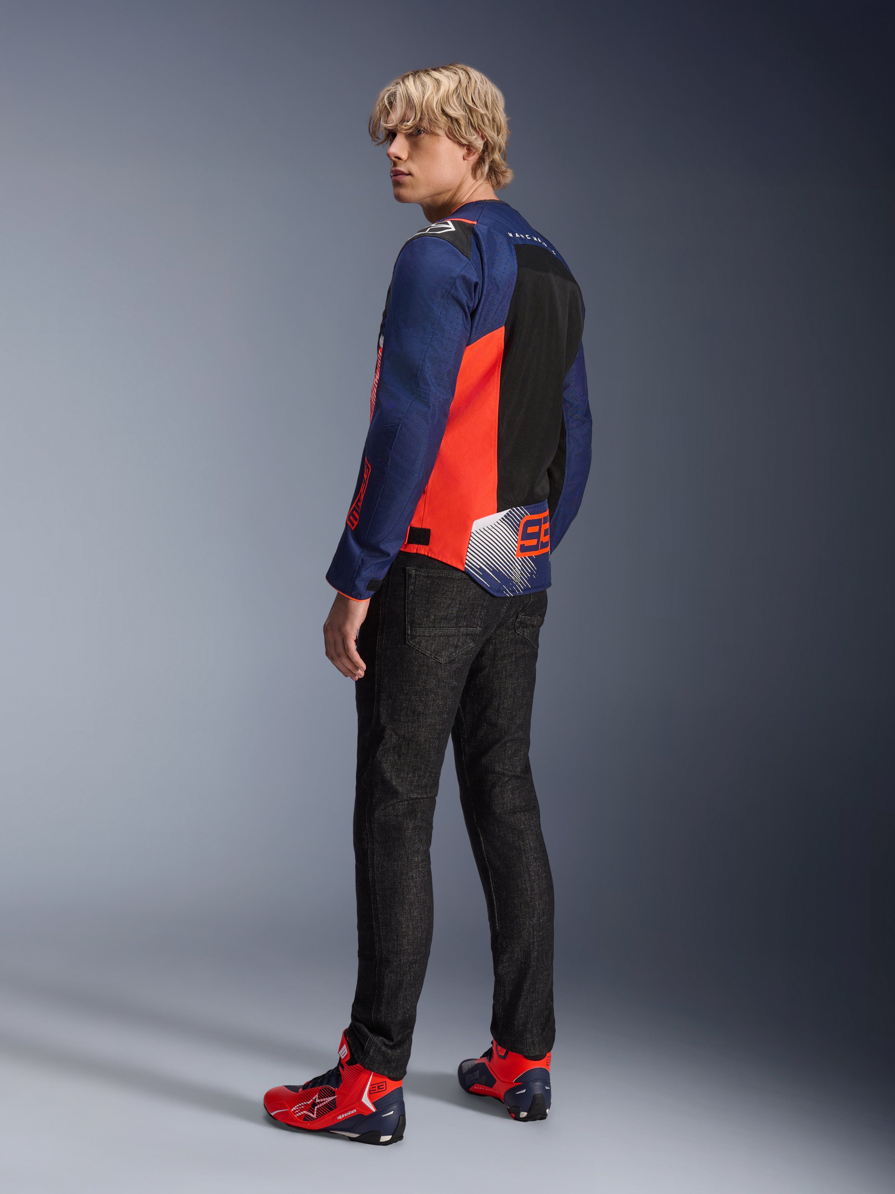 Alpinestars MM93 T-SPS Air V2 Jacket, Blue Navy and Red Fluo, worn by a model from the back with black riding jeans and matching red and blue shoes, featuring mesh panels and Marc Márquez 93 branding.