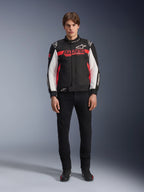 Male model wearing the Alpinestars Monza Sport Jacket in Black, Bright Red, and White, featuring a large red 'ASTARS' logo across the chest and white panels on the sleeves, paired with black pants and black riding boots.