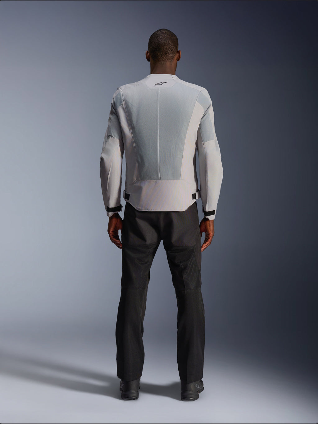 Alpinestars C-1 Air Jacket in Silver, viewed from the back on a male model, textile motorcycle jacket featuring large tech mesh ventilation panels, small black logo at the neck, and waist adjusters, worn with black riding pants for an urban contemporary look.