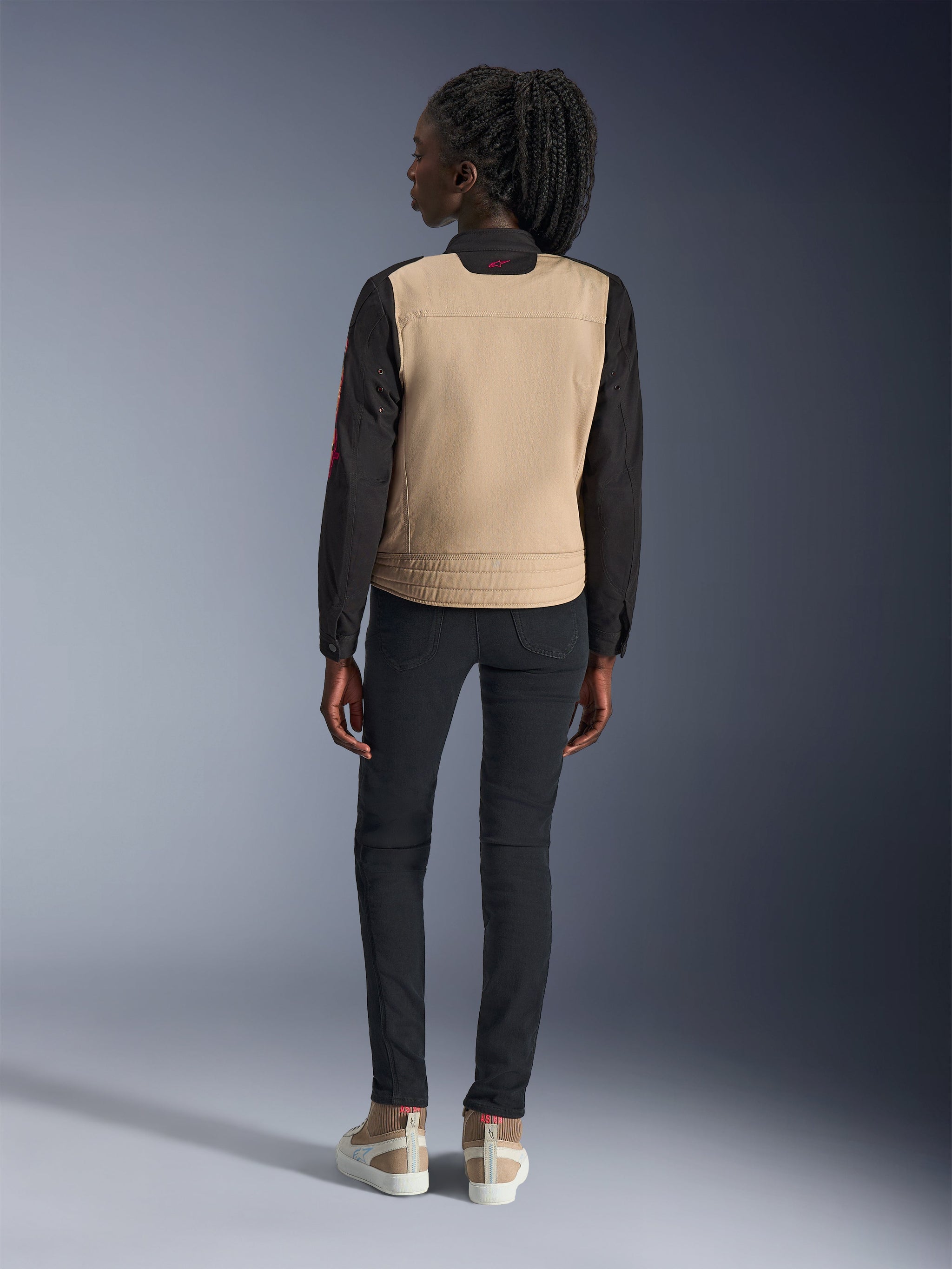 Woman wearing Alpinestars Stella Grace Jacket, Aluminum Black with tan torso and black sleeves, rear view, styled with black slim-fit pants and casual sneakers, urban motorcycle gear for women