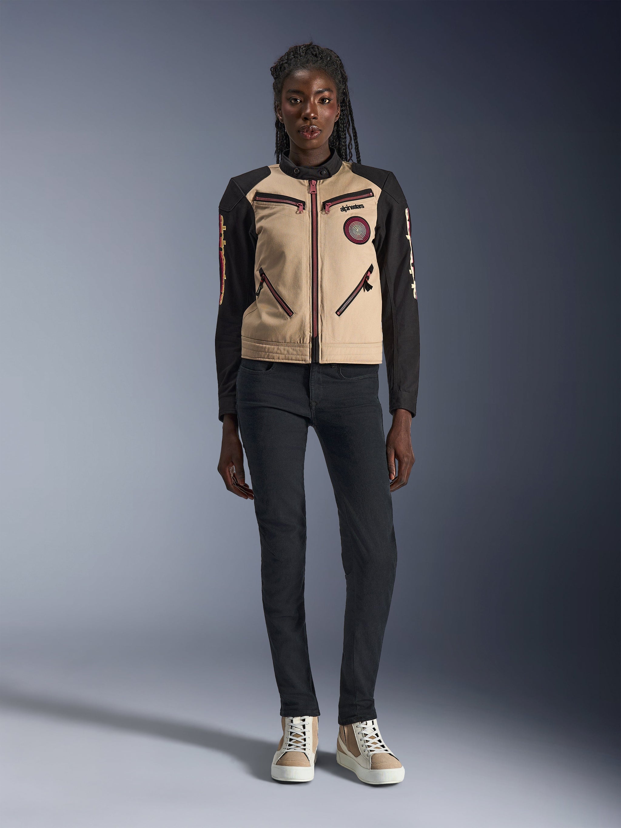 Alpinestars Woman Stella Grace Jacket, urban motorcycle jacket, tan and black textile construction, featuring front zippers and embroidered Oscar spiral patch, worn with black pants and high-top sneakers, contemporary workwear-inspired riding gear