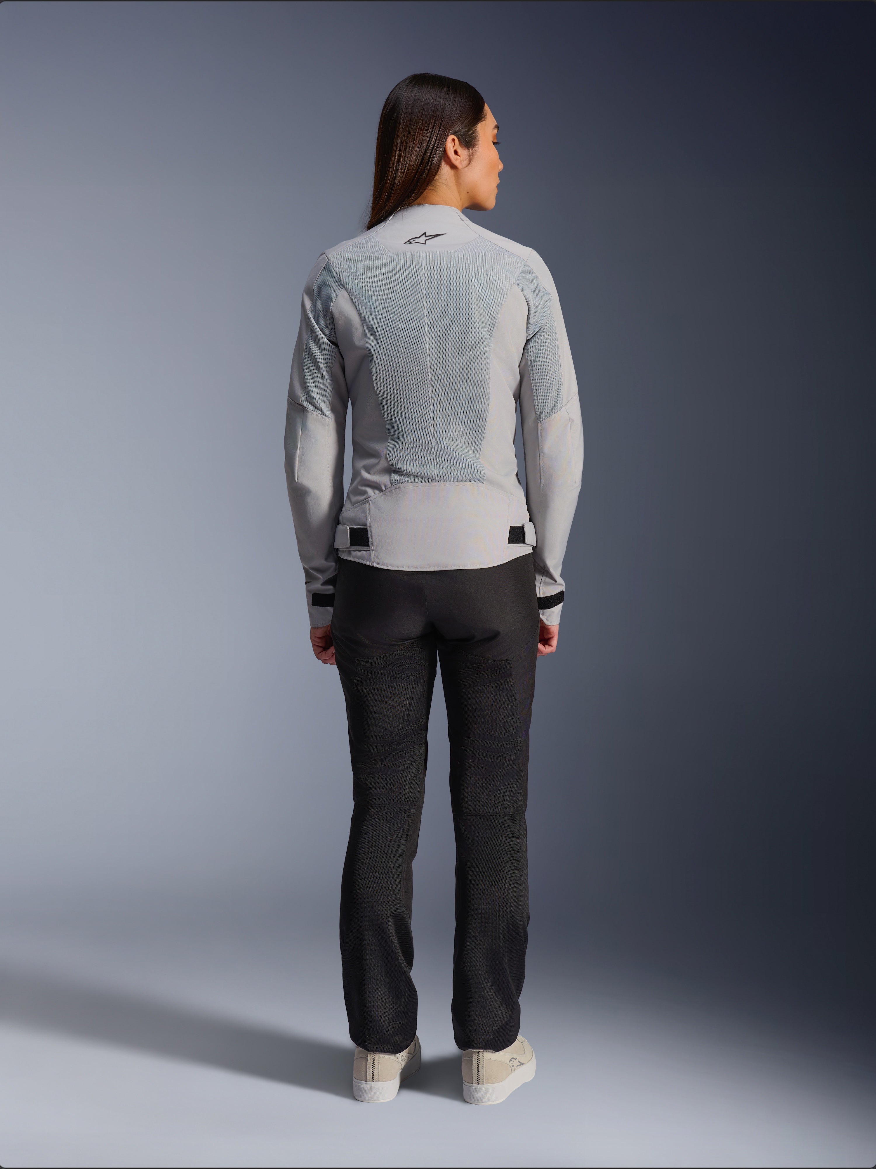 Woman wearing Alpinestars Stella C-1 Air Jacket in Silver, back view, featuring large tech mesh panels for ventilation and a reflective star logo, paired with black riding pants and white sneakers, urban contemporary motorcycle gear