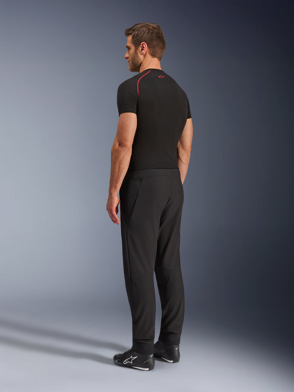 Alpinestars Aeroshell Airflow Pants, Black motorcycle pants, lightweight softshell textile, worn with a black technical short-sleeve shirt and black riding boots, rear-side profile view showing the slim fit and ergonomic construction.