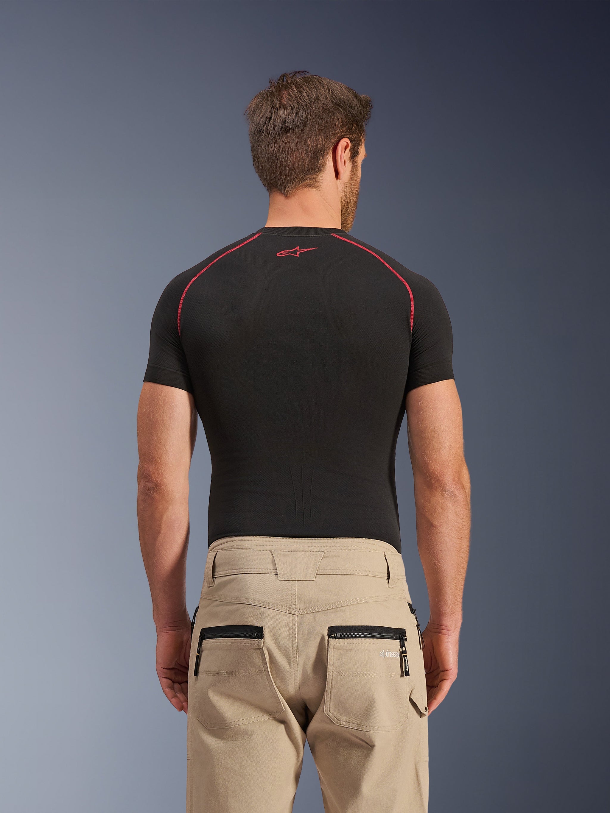Alpinestars Flex-AST Canvas Pants in brown, rear view showing zippered back pockets and workwear-inspired hammer loop, worn with a black short-sleeve base layer featuring red contrast stitching, urban motorcycle riding apparel
