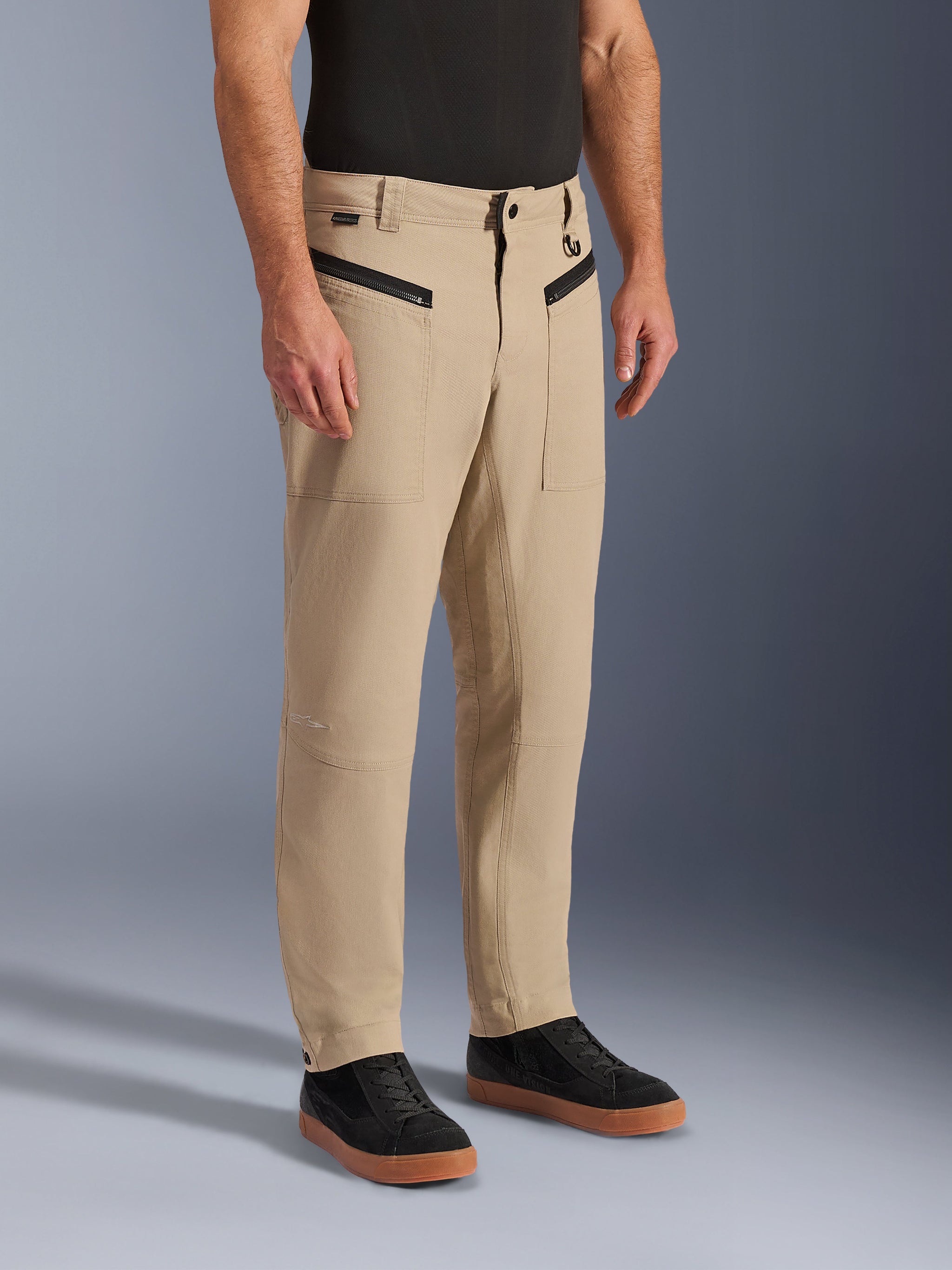 Alpinestars Flex-AST Canvas Pants, urban motorcycle pants in tan brown, monolayer stretch polycotton with workwear styling, featuring zippered front pockets and tapered legs, worn with a black t-shirt and black sneakers, casual protective gear for city riding