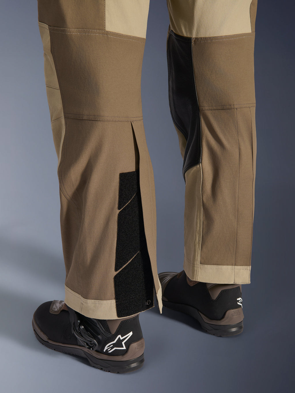 Alpinestars Maxdura Dual Pants in Walnut Aluminum, detailed view of the lower leg and hem featuring an over-the-boot design with adjustable hook-and-loop closures and reinforced inner panels, paired with black motorcycle boots.