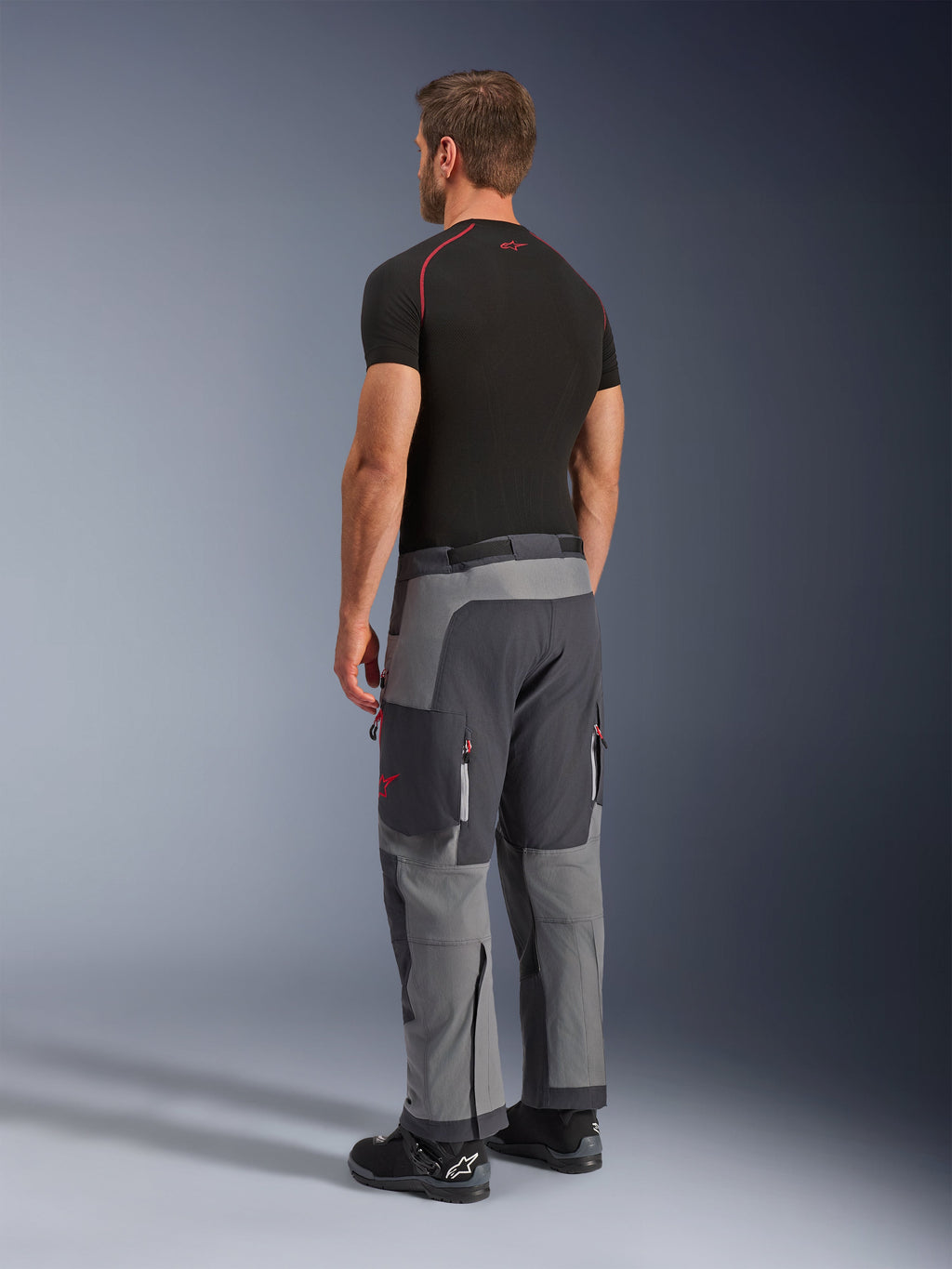 Alpinestars Maxdura Dual Pants, Adventure Motorcycle Pants, Dark Gray and Deep Gray, worn by a model from a back-side view, featuring red accents on zippers and logo, reinforced gray panels, cargo pockets, and over-the-boot fit