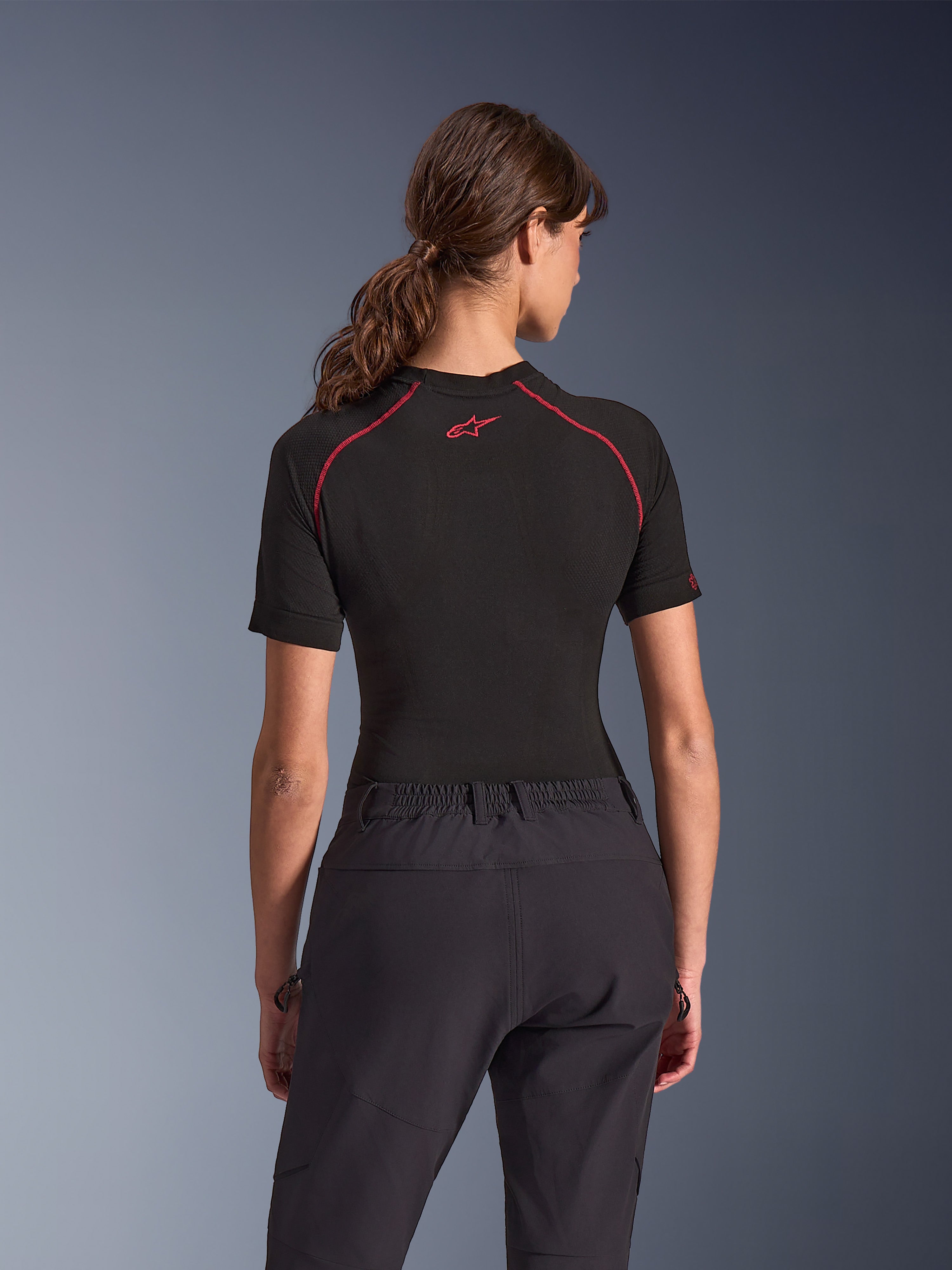 Woman wearing Alpinestars Stella Flex-AST Explorer Pants, Black adventure motorcycle pants, back view, paired with a black short-sleeved top with red stitching, highlighting the breathable textile construction and elastic waistband.