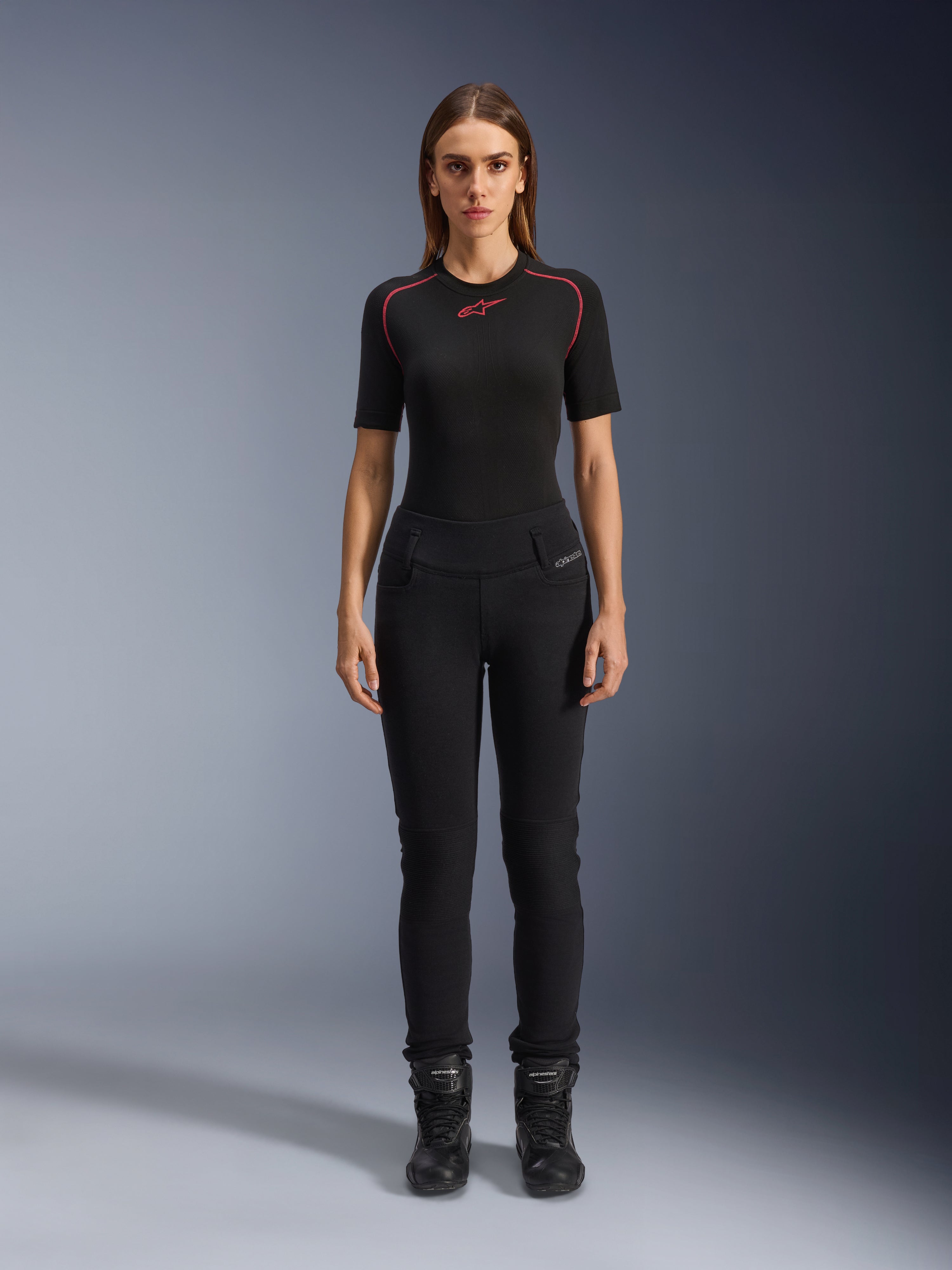 Alpinestars Womens Banshee Leggings, Black motorcycle leggings, figure-hugging anatomical fit, worn by a female model with a black base layer and motorcycle boots, featuring ribbed knee panels and high-waisted construction for urban riding.