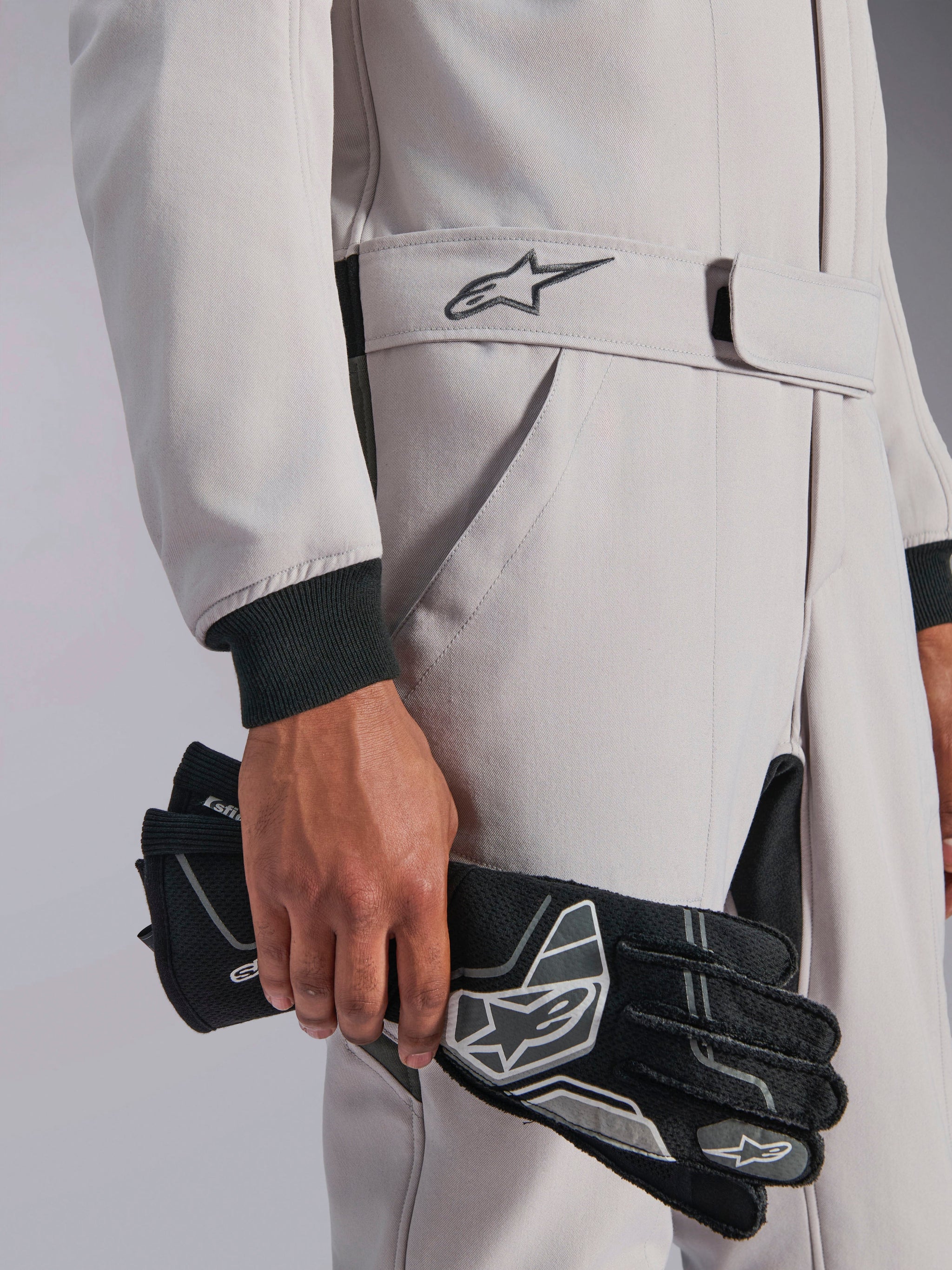 Alpinestars GP Race V3 Suit FIA, Auto Racing Suit, Silver Anthracite, held with black racing gloves, featuring adjustable belted waist with embroidered star logo, black ribbed cuffs, clean and professional motorsport design