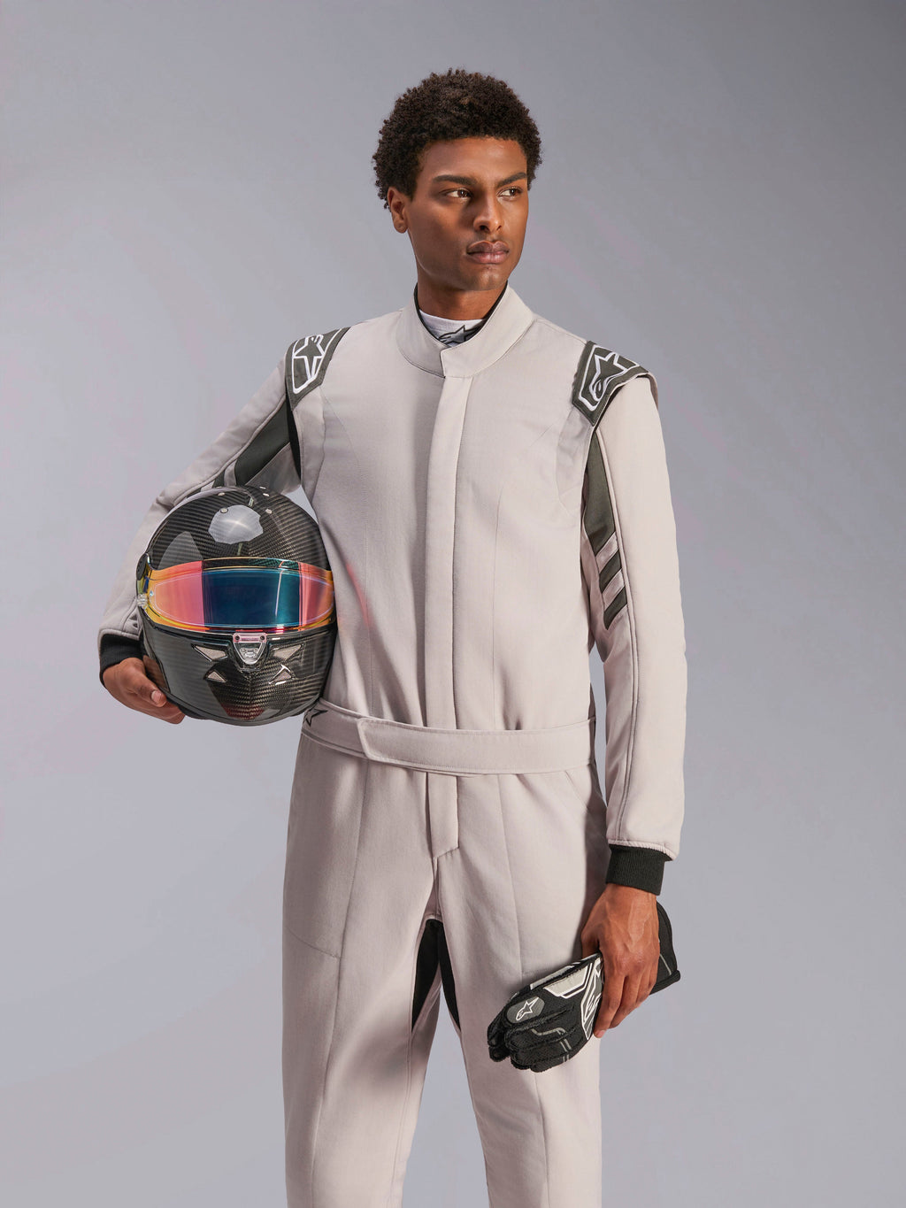 Alpinestars GP Race V3 Suit FIA, Auto Racing Suit, Silver Anthracite, worn by model holding a carbon fiber helmet with an iridescent visor and black racing gloves, professional motorsport protective gear with visible shoulder epaulettes.