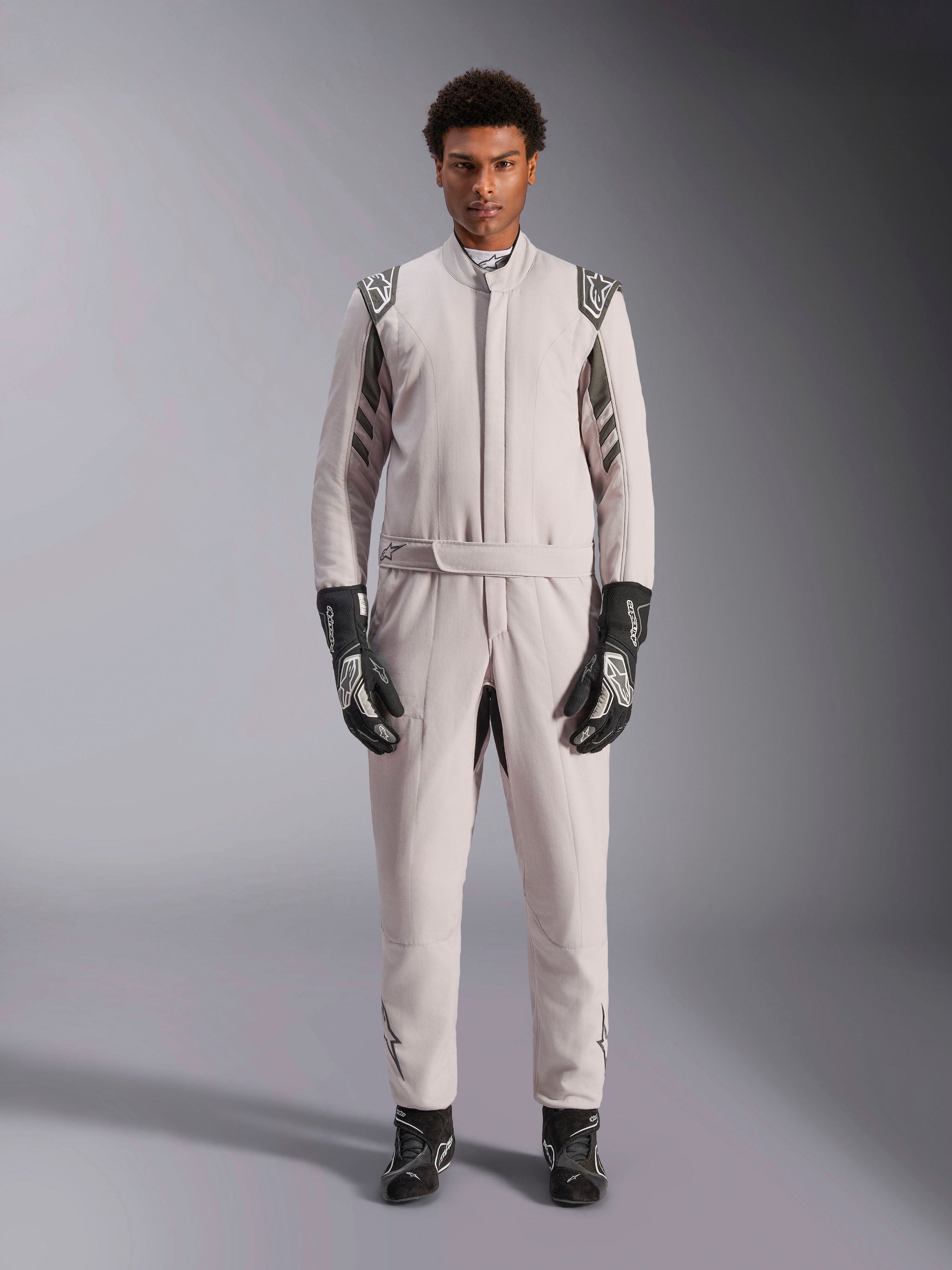 Alpinestars GP Race V3 Suit FIA in Silver Anthracite, one-piece auto racing suit worn by a model, featuring dark gray epaulettes and accents, paired with black racing gloves and boots, professional motorsport protective gear.