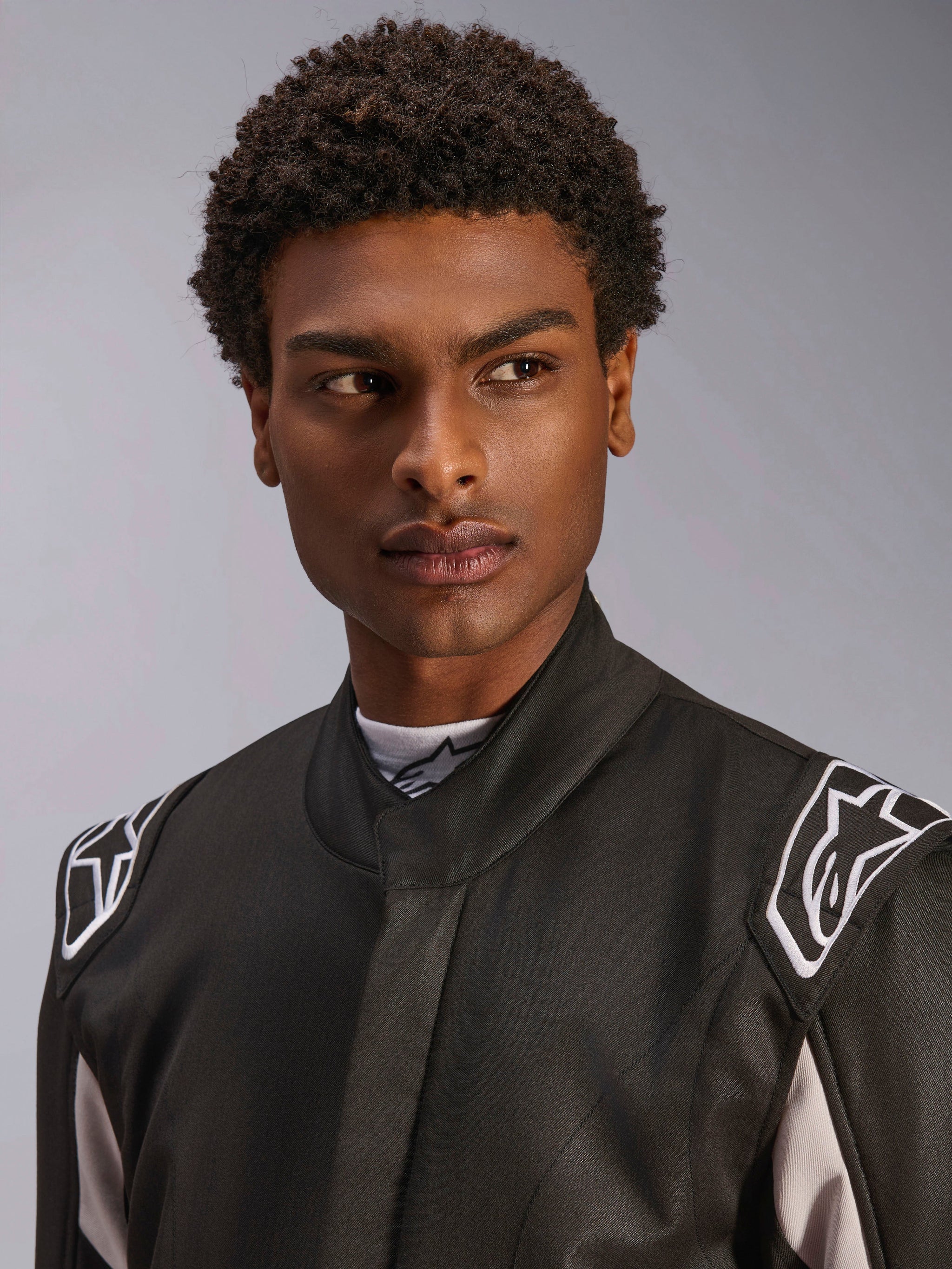Alpinestars GP Race V3 Suit FIA, Black auto racing suit worn by a man, close-up view showing the soft collar and white star logos on the shoulder epaulettes, professional motorsport protective gear.
