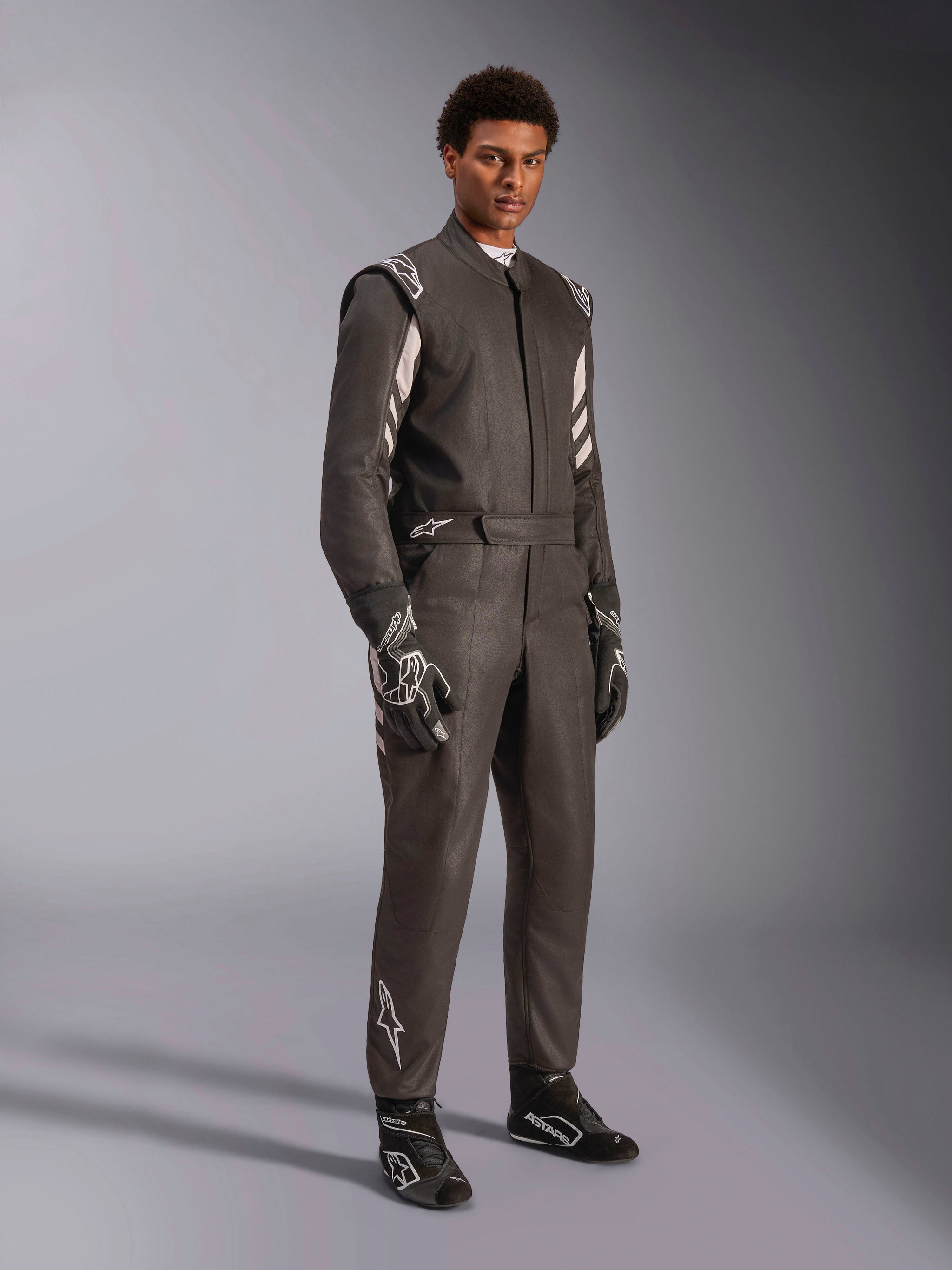 Male racer wearing Alpinestars GP Race V3 Suit FIA, Black and Silver auto racing suit, complete with matching gloves and shoes, featuring silver accents on the shoulders and lower leg, professional automotive racing gear with a tailored fit