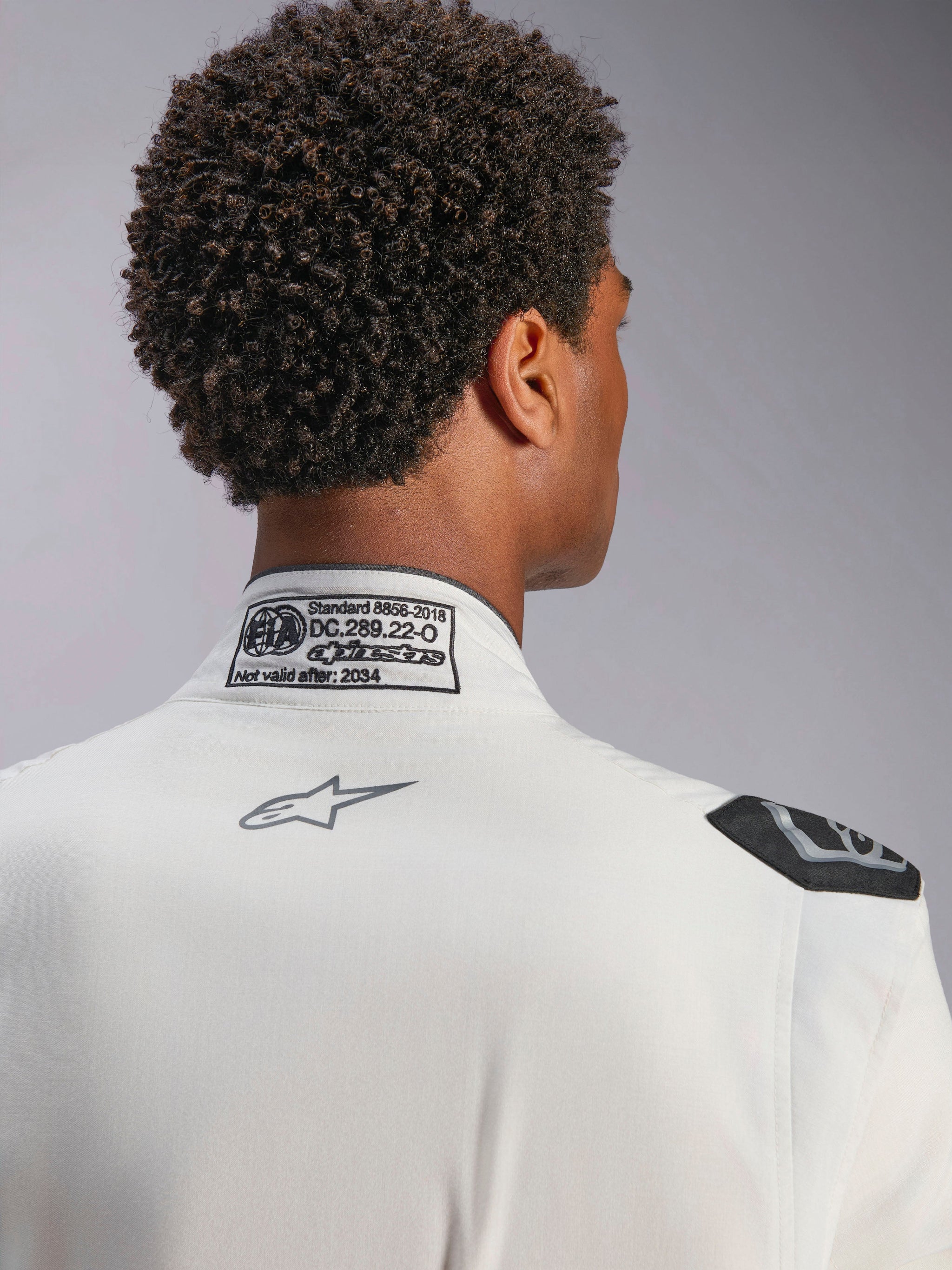 Alpinestars Hypertech V3 Suit, White, detail view of back collar and shoulder, featuring FIA 8856-2018 certification label, printed grey Alpinestars logo, and black extraction handle, professional racing gear