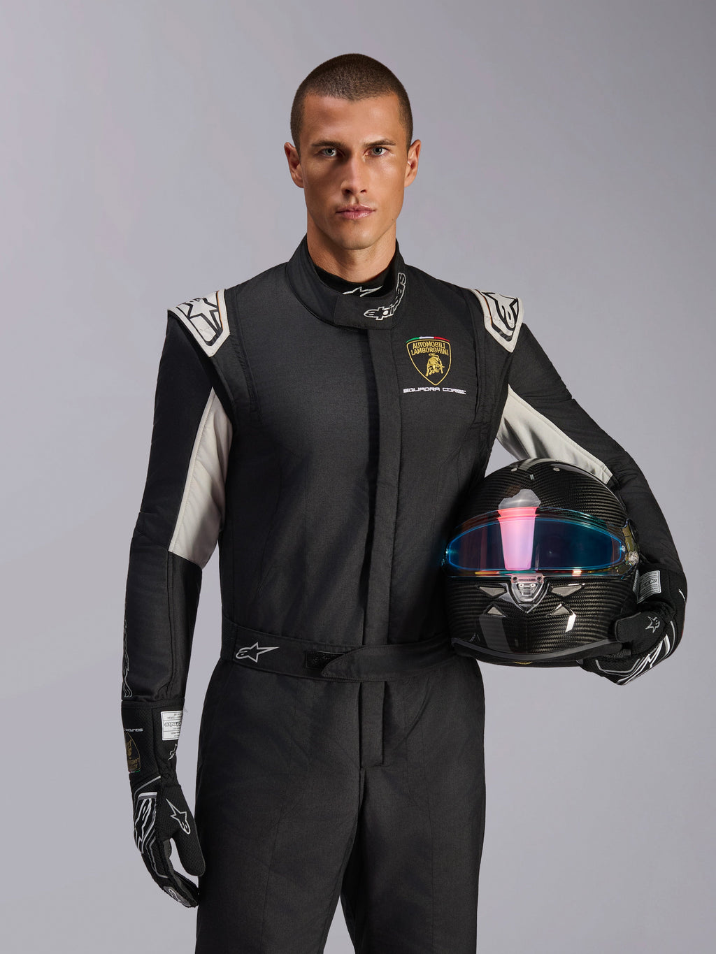 Alpinestars Limited Edition Lamborghini GP Tech V4 Suit, Auto Racing Suit, Black Silver and Gold, worn by a model holding a carbon fiber helmet with an iridescent visor, featuring Lamborghini Squadra Corse branding and white shoulder accents