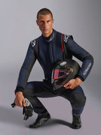 Male racing driver wearing Alpinestars GP Tech V4 Suit, Navy Blue with black and red accents, holding a carbon fiber helmet with an iridescent visor, black racing gloves and boots, professional automotive racing gear