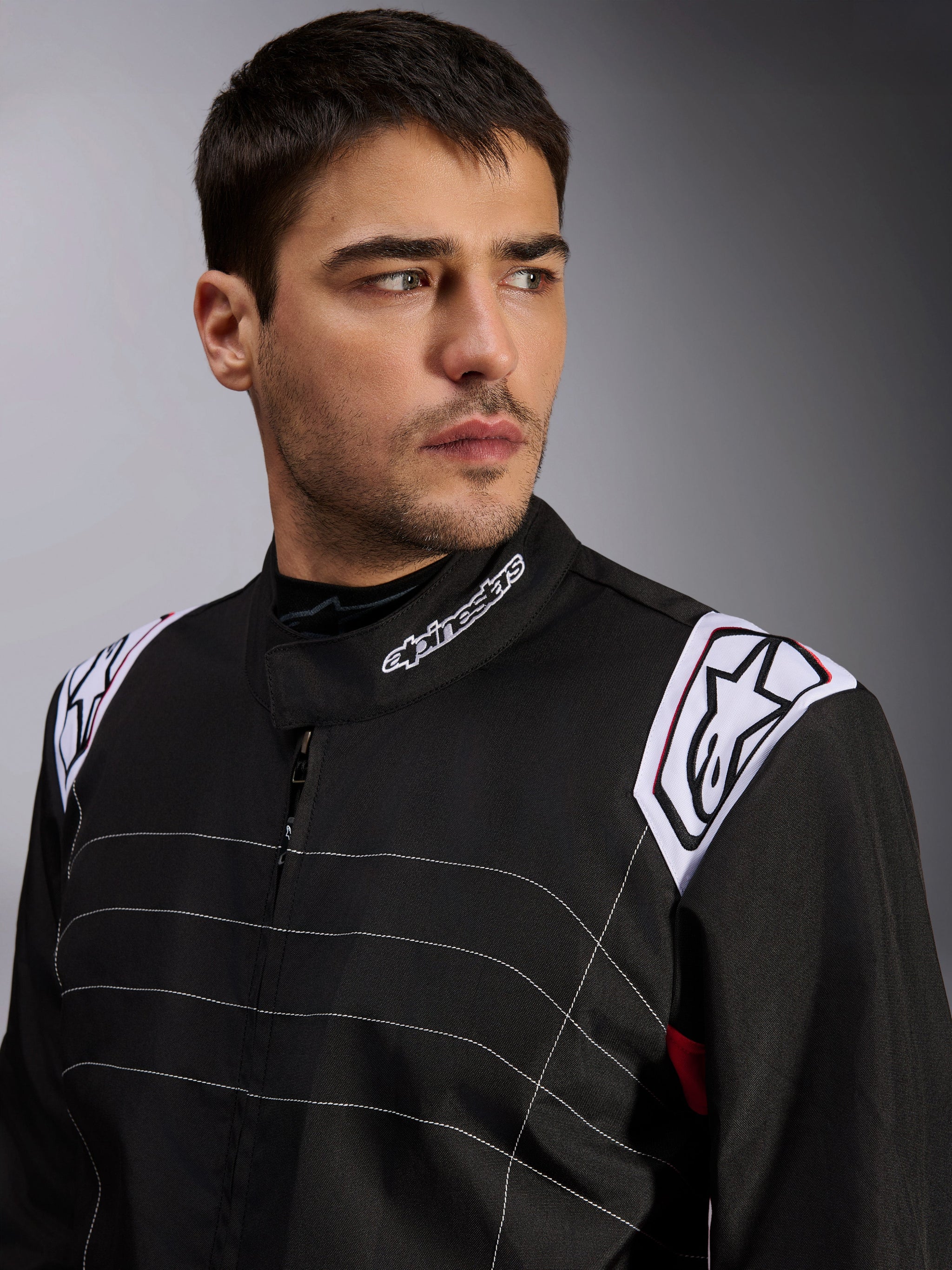 Alpinestars KMX-9 V3 Suit, Black Karting Suit worn by a male model, close-up of chest and shoulders with white branding on the collar and epaulettes, featuring white decorative stitching and a professional motorsport design