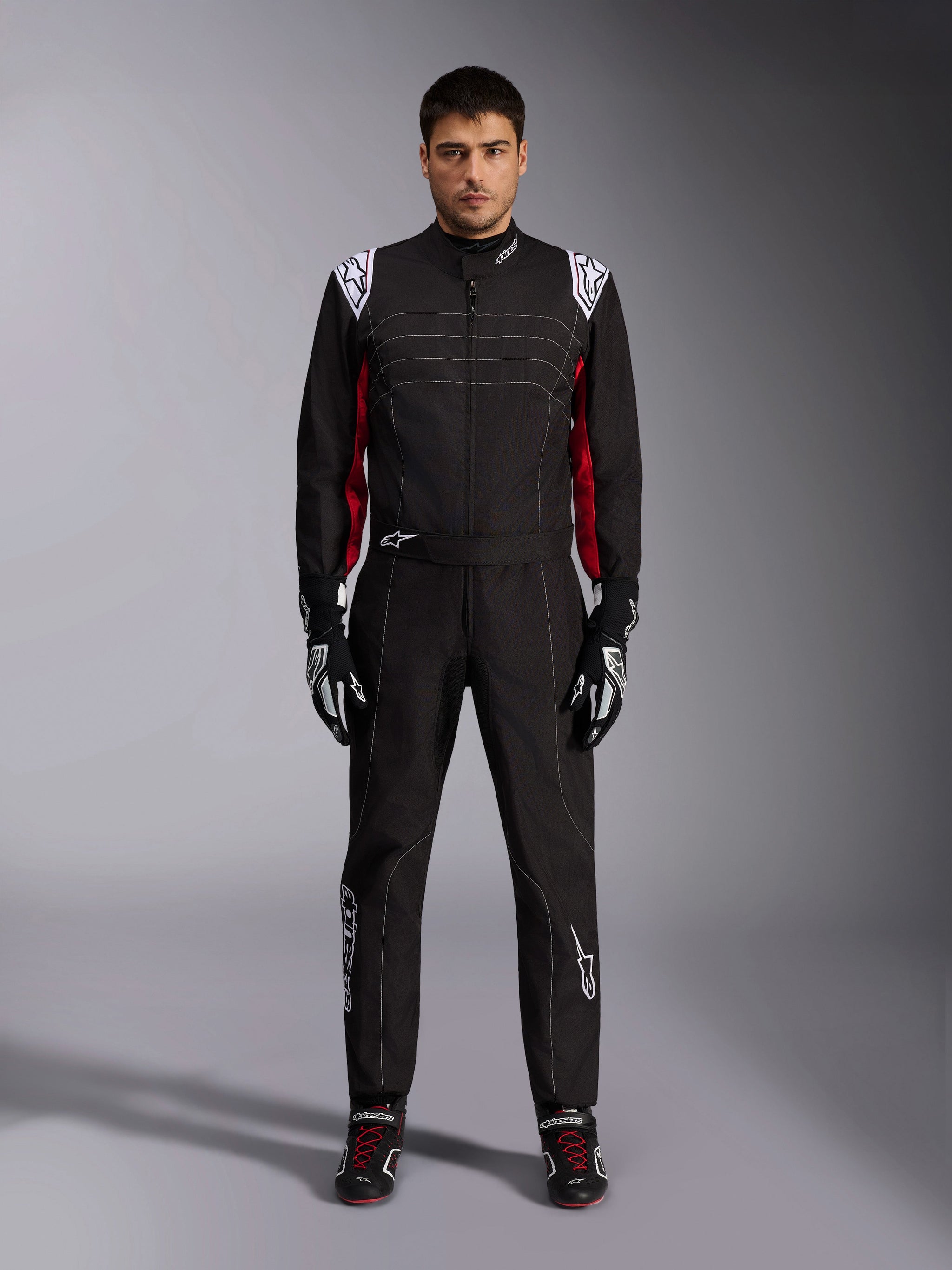 Alpinestars KMX-9 V3 Karting Suit in Black Red White, full-body front view of a model wearing the one-piece suit with coordinated racing gloves and shoes, featuring white Alpinestars logos on the shoulders and red accents.