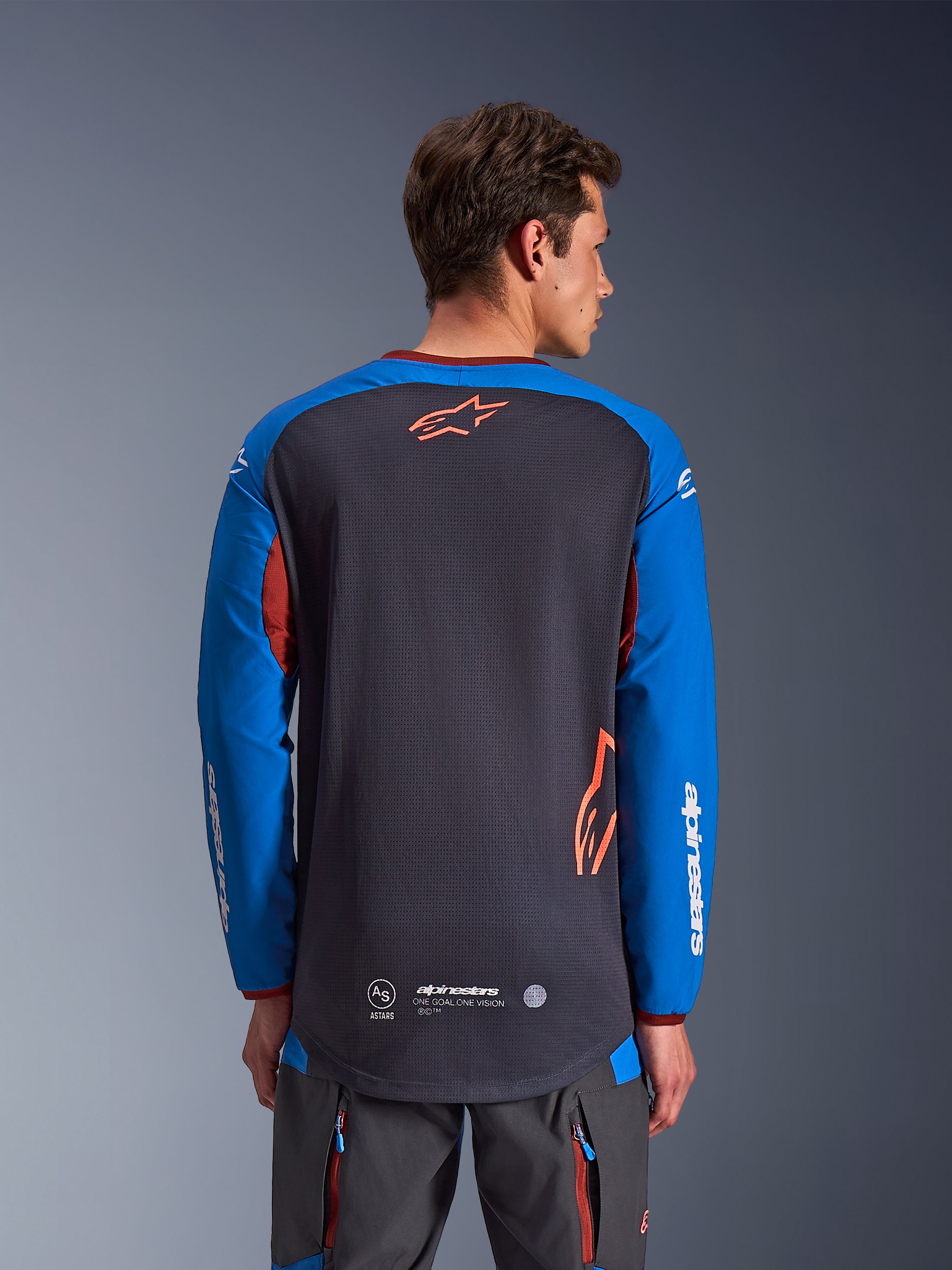 Alpinestars Maxdura Dual Jersey in Deep Gray and Royal Blue, back view of male model, mesh torso construction with orange Astars logo, long sleeves with white branding, maroon collar and cuffs, worn with gray technical pants, dual-sport and adventure motorcycle gear