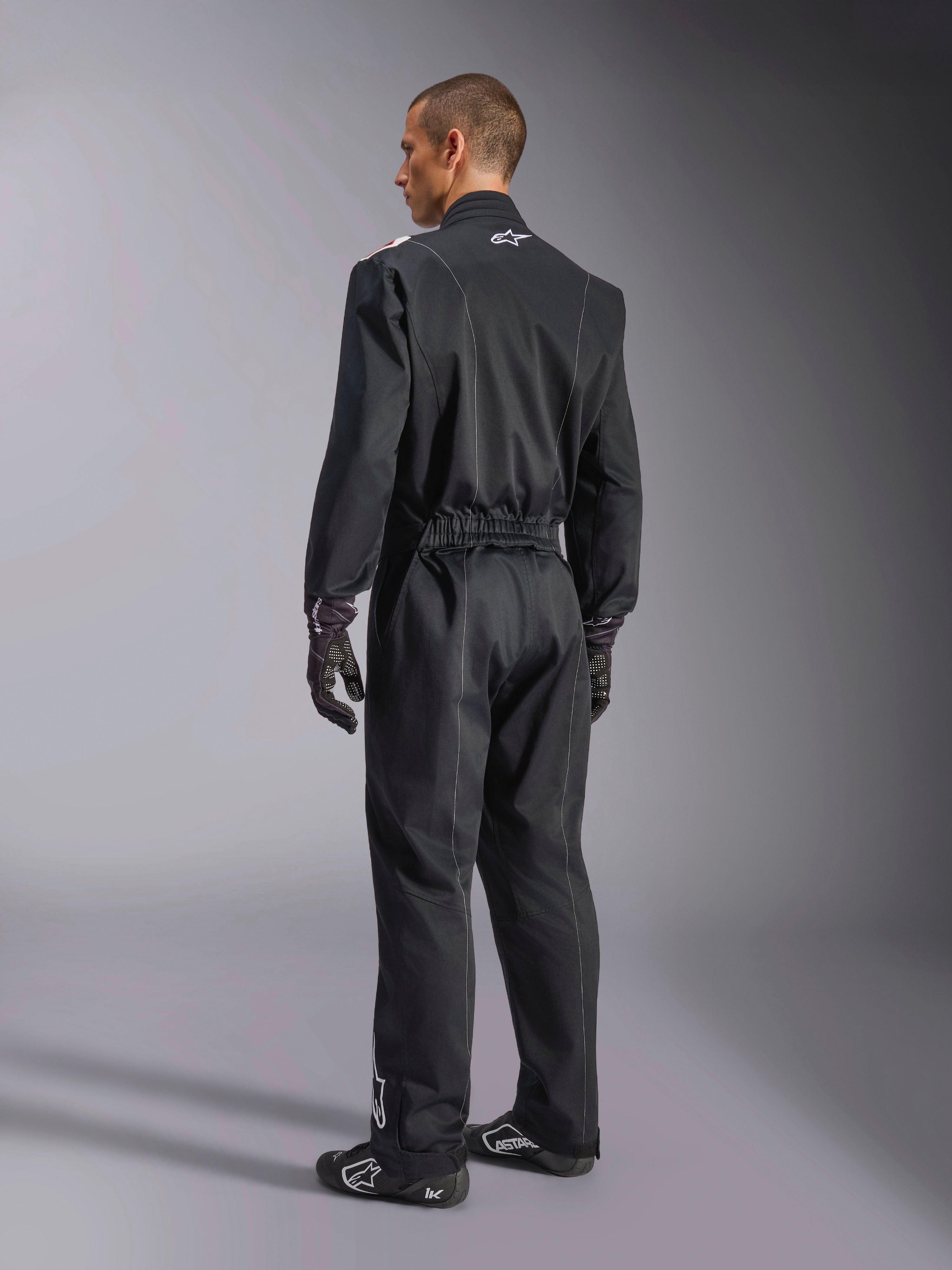Alpinestars Kart Indoor V2 Suit, Black one-piece karting suit shown from a back three-quarter angle, featuring an elastic waist and a white logo on the neck, worn with black racing gloves and shoes, durable design for recreational karting