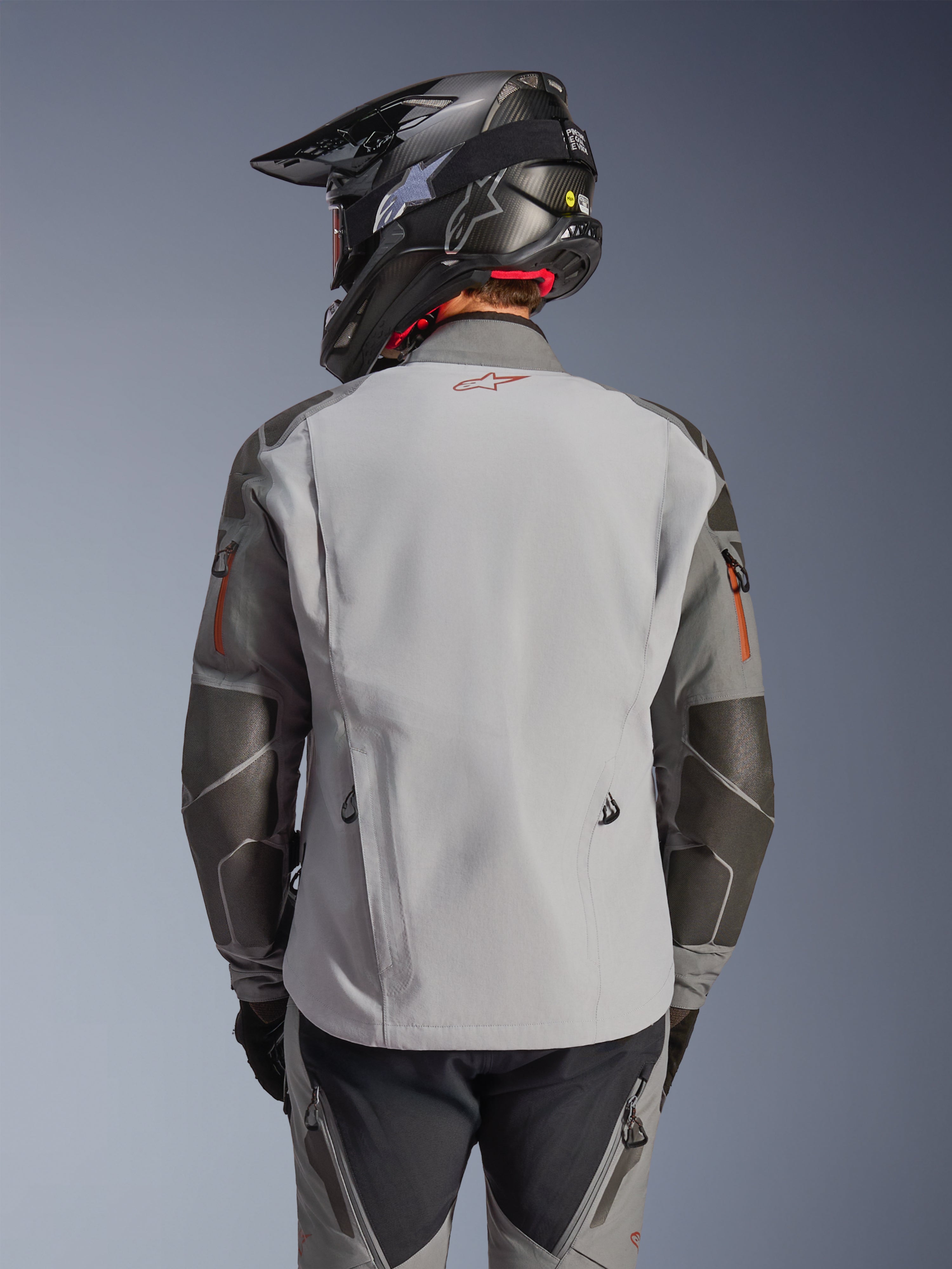 Alpinestars Nazca 3L GORE-TEX PRO Jacket, Adventure Motorcycle Jacket, Back View, Dark Gray and Mid Gray, features Superfabric reinforcements on elbows and shoulders, red logo on the back, worn with matching adventure gear and a carbon fiber helmet.