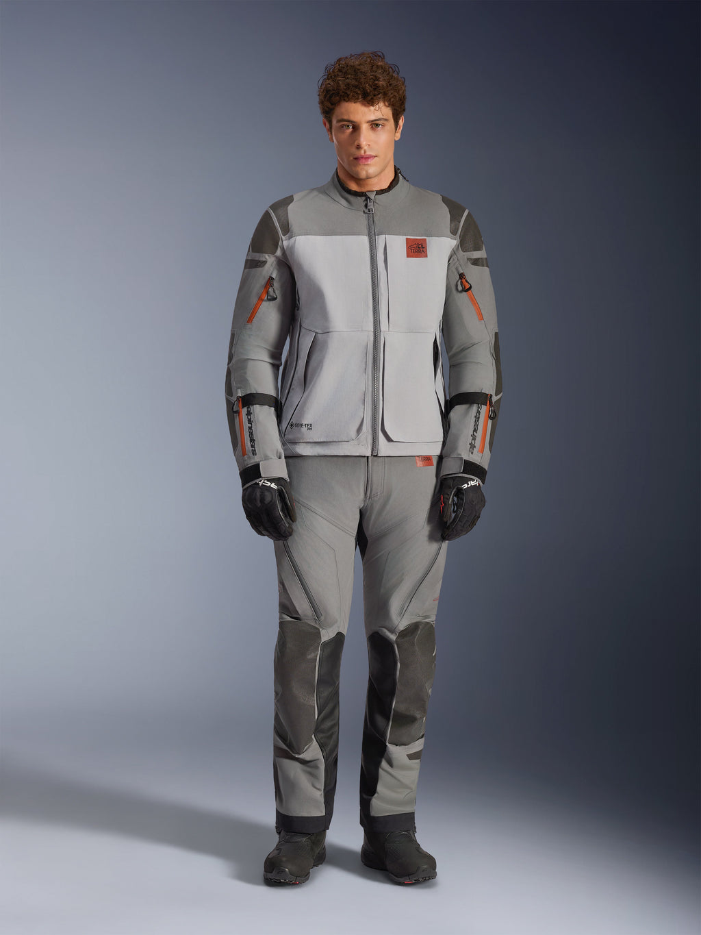 Alpinestars Nazca 3L GORE-TEX PRO Jacket, Adventure Motorcycle Jacket, Dark Gray and Mid Gray, worn by a model with matching pants and black gloves, featuring orange zipper accents and red chest logo, technical protective gear for extreme riding