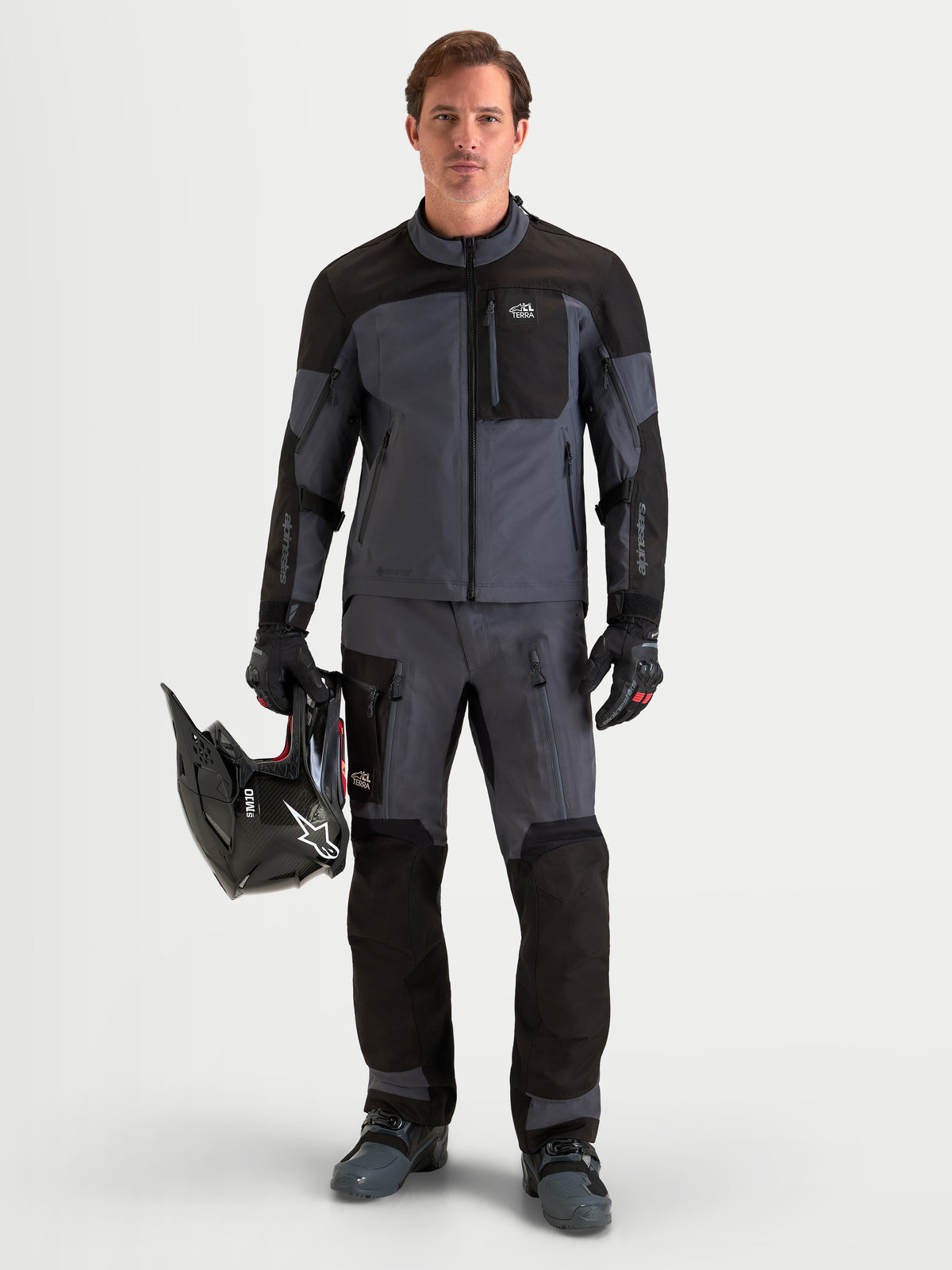 Alpinestars Arenis Gore-Tex Jacket, men's adventure touring motorcycle jacket in black and deep gray, worn by a rider holding a helmet, showcasing waterproof GORE-TEX textile construction.
