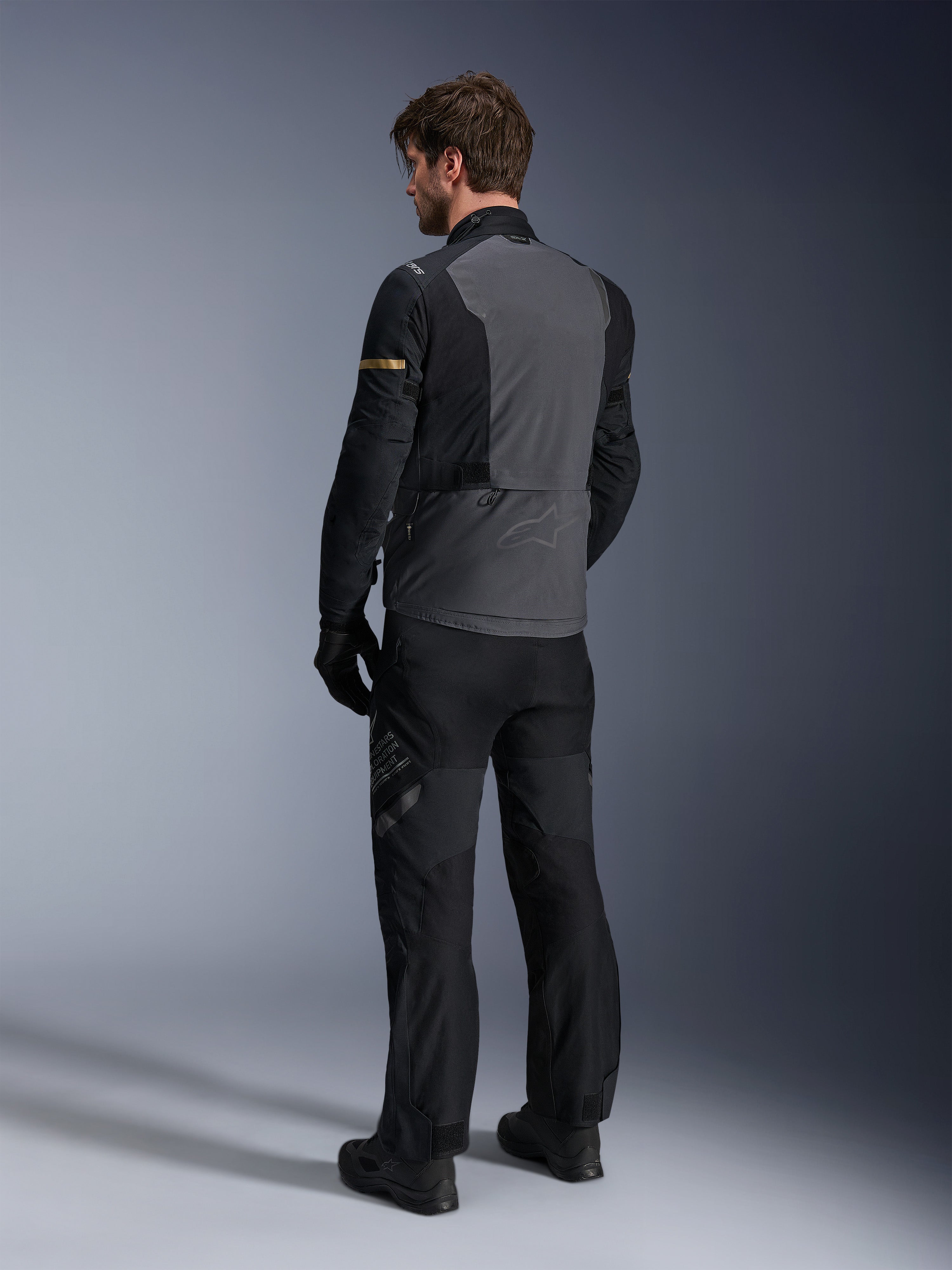 Alpinestars ST-7 2L GORE-TEX Jacket, Deep Gray and Black with Dark Khaki accents, rear view of a rider wearing the jacket and matching motorcycle pants, adventure touring gear with waterproof laminated construction