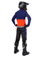 Motocross rider from the back wearing Alpinestars Lite-Dura Softshell Jacket in Navy, Hot Orange, and White, showing the large rear storage pocket. Styled with matching enduro pants, a black motocross helmet, and black off-road boots.