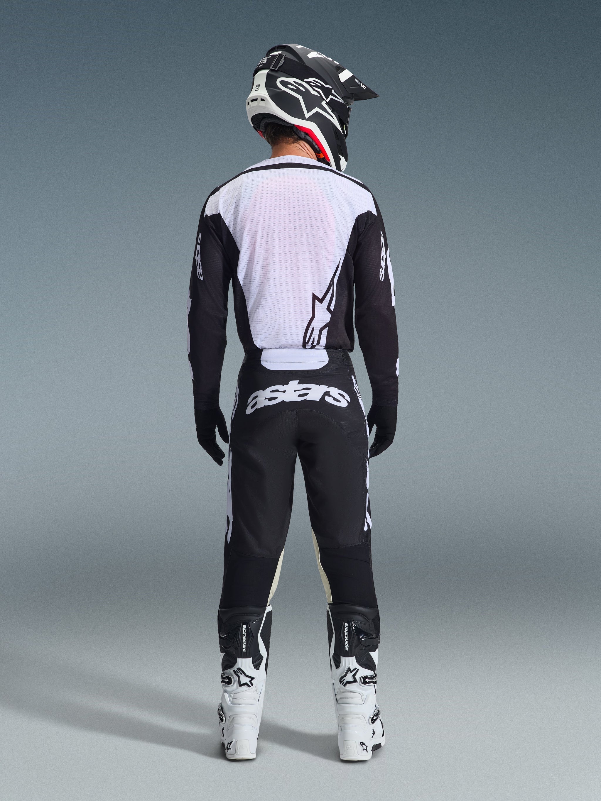 Motocross rider seen from behind wearing Alpinestars 2026 Racer Air Riway Pants in Black and White, matching black and white long-sleeve jersey, white and black racing boots, and a black helmet with Alpinestars branding