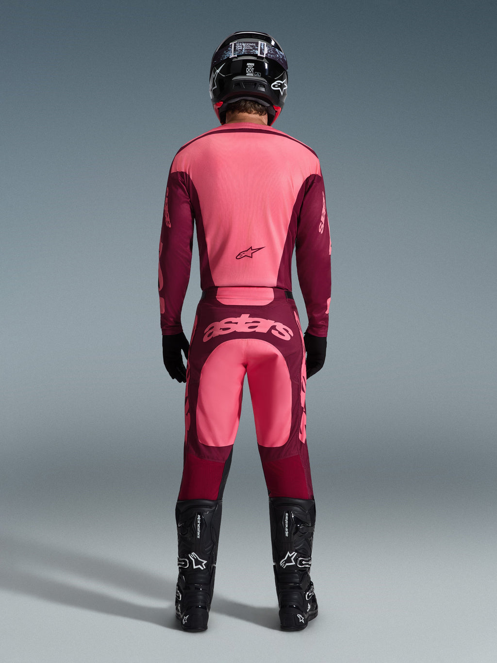 Rear view of a motocross rider wearing the 2026 Racer Riway Gear Set, pink and burgundy jersey, matching burgundy pants with light pink 'astars' logo on the seat, black helmet, black boots, and black gloves, coordinated outfit for off-road performance