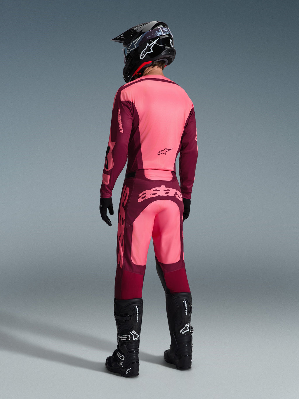 Motocross rider wearing the Alpinestars 2026 Racer Riway Gear Set from a rear view, maroon and light pink colorway, jersey and pants with prominent branding, complete with black helmet, gloves, and boots, coordinated motocross racing outfit
