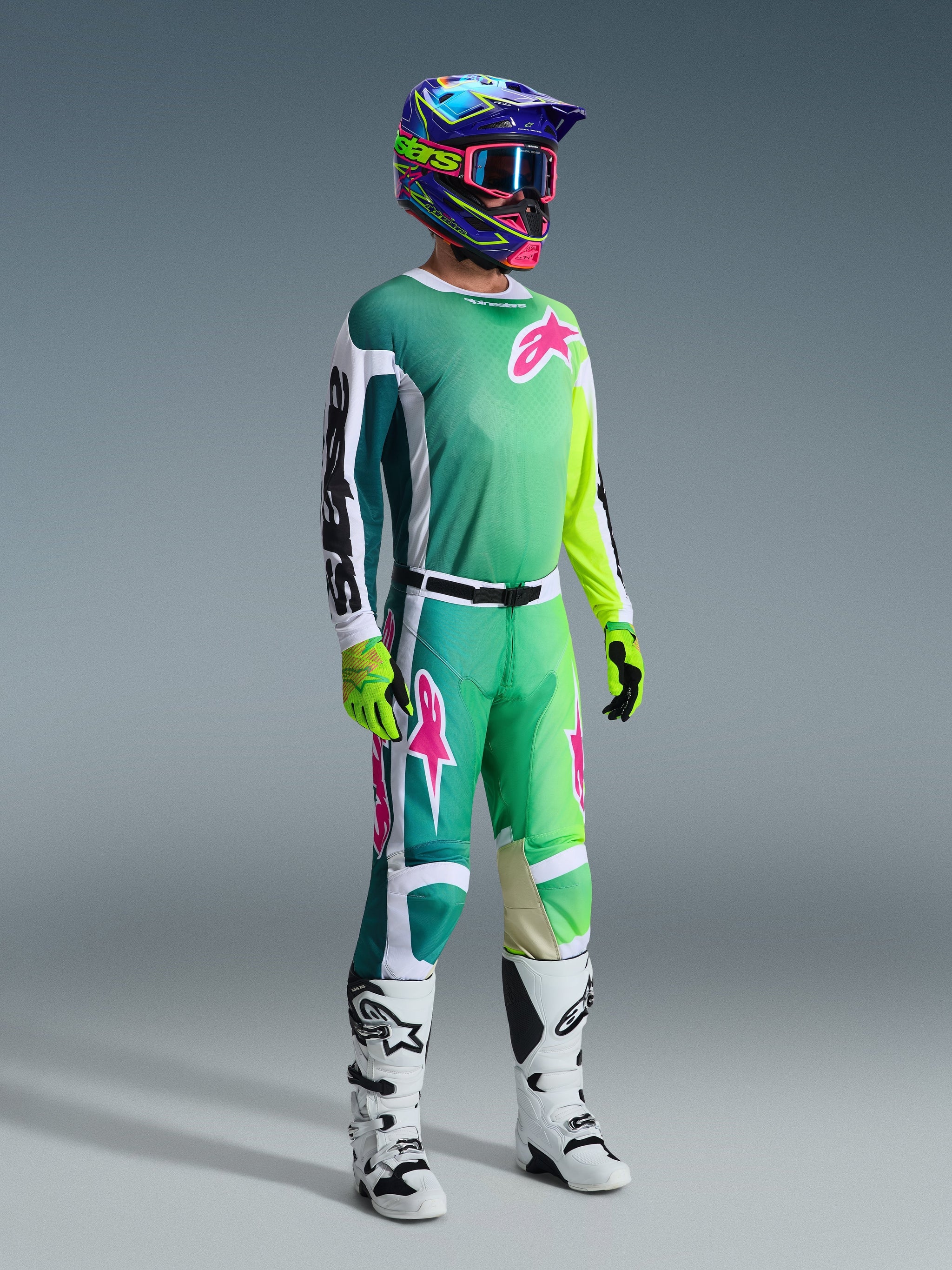 Motocross rider wearing Alpinestars 2026 Racer Portl Pants and matching jersey in Green, White, and Black with pink logo accents, colorful helmet, white and black racing boots, and neon green gloves, complete gear set for off-road racing
