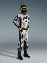 Motocross rider wearing full Alpinestars racing gear, 2026 Racer Lahnd Pants in Sand Camo, matching camo jersey with bold branding, black helmet with neon yellow goggles, neon green gloves, black boots, coordinated professional outfit for off-road performance.