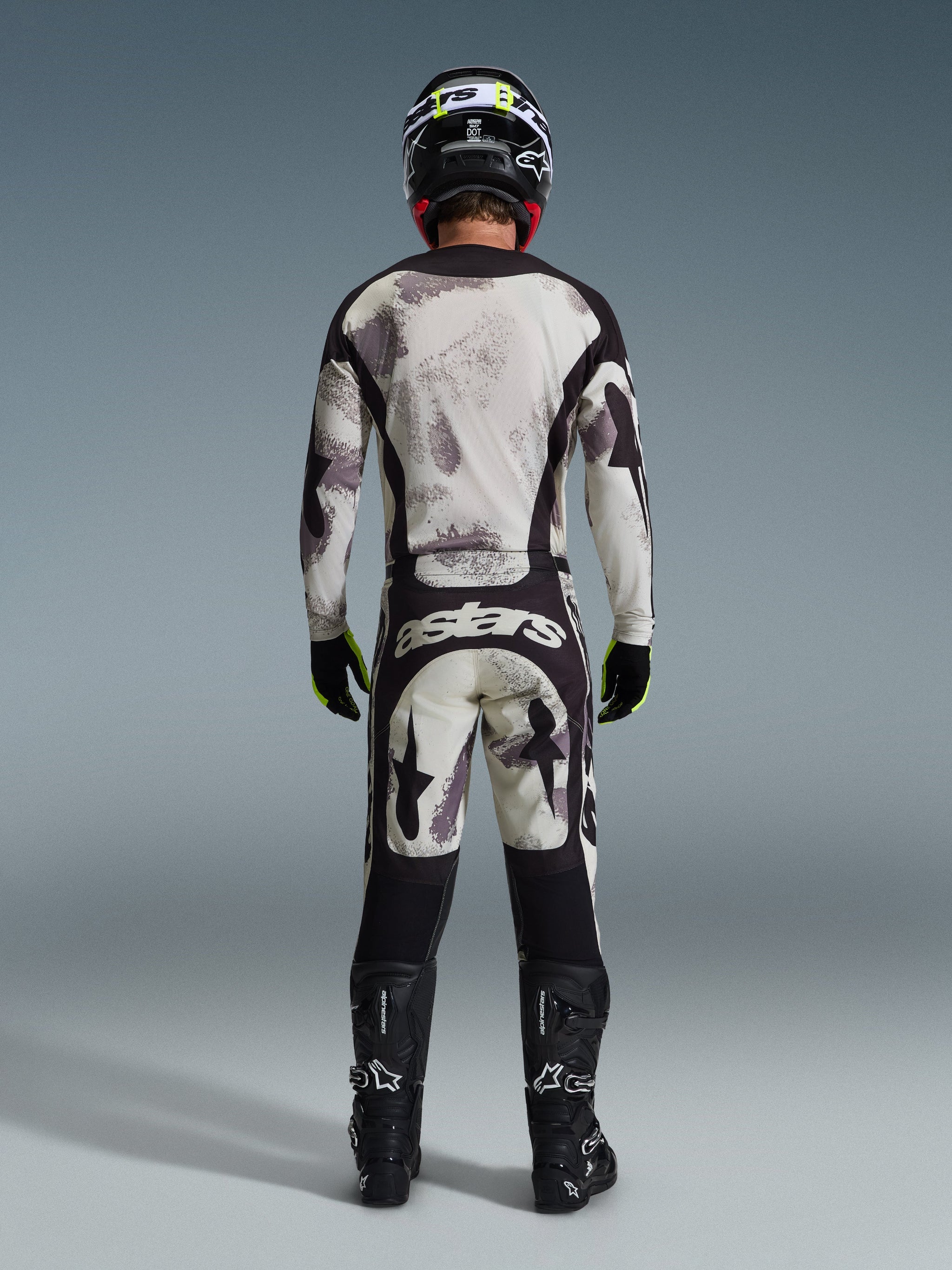 Motocross rider from behind wearing the Alpinestars 2026 Racer Lahnd Pants in Sand Camo, matching long-sleeve jersey with gray camouflage pattern, black motocross boots, black helmet, and "astars" branding on the seat, designed for professional off-road racing.