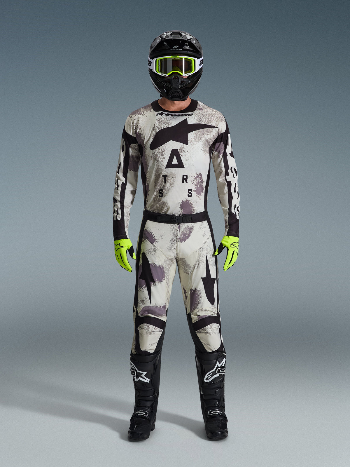 Motocross rider wearing the Alpinestars 2026 Racer Lahnd Gear Set, Sand Camo patterned suit with white, gray, and black design, black helmet with neon green goggles, bright neon green gloves, and black boots with white logos, complete coordinated outfit for off-road racing
