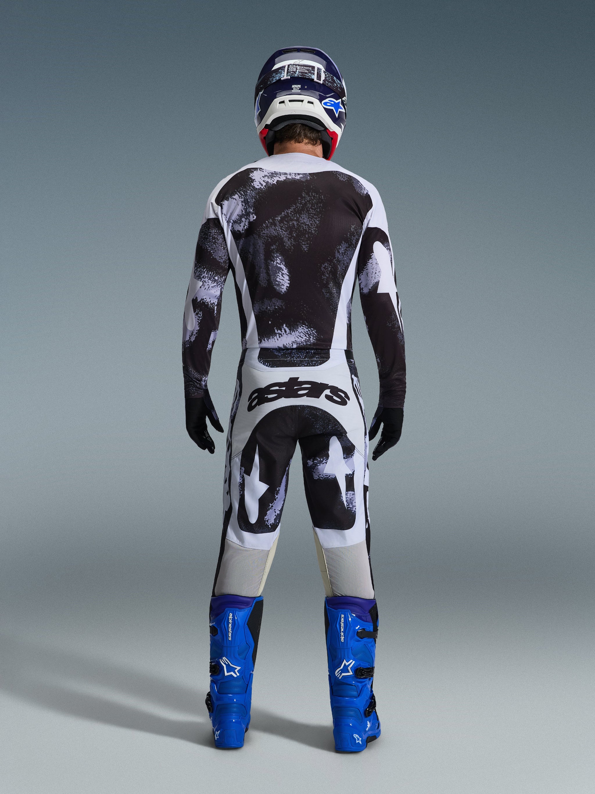 Alpinestars 2026 Racer Lahnd Pants, Motocross Pants, Iron Camo gray and black pattern, back view of rider in full racing gear with matching jersey and blue boots, designed for off-road performance.
