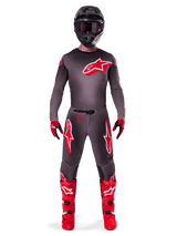 Motocross rider wearing complete Alpinestars 2025 Supertech Lipan gear set, Smoke Bright Red, featuring a dark grey jersey and pants with prominent red logos, black helmet with goggles, red and black gloves, and vibrant red motocross boots, coordinated professional racing outfit