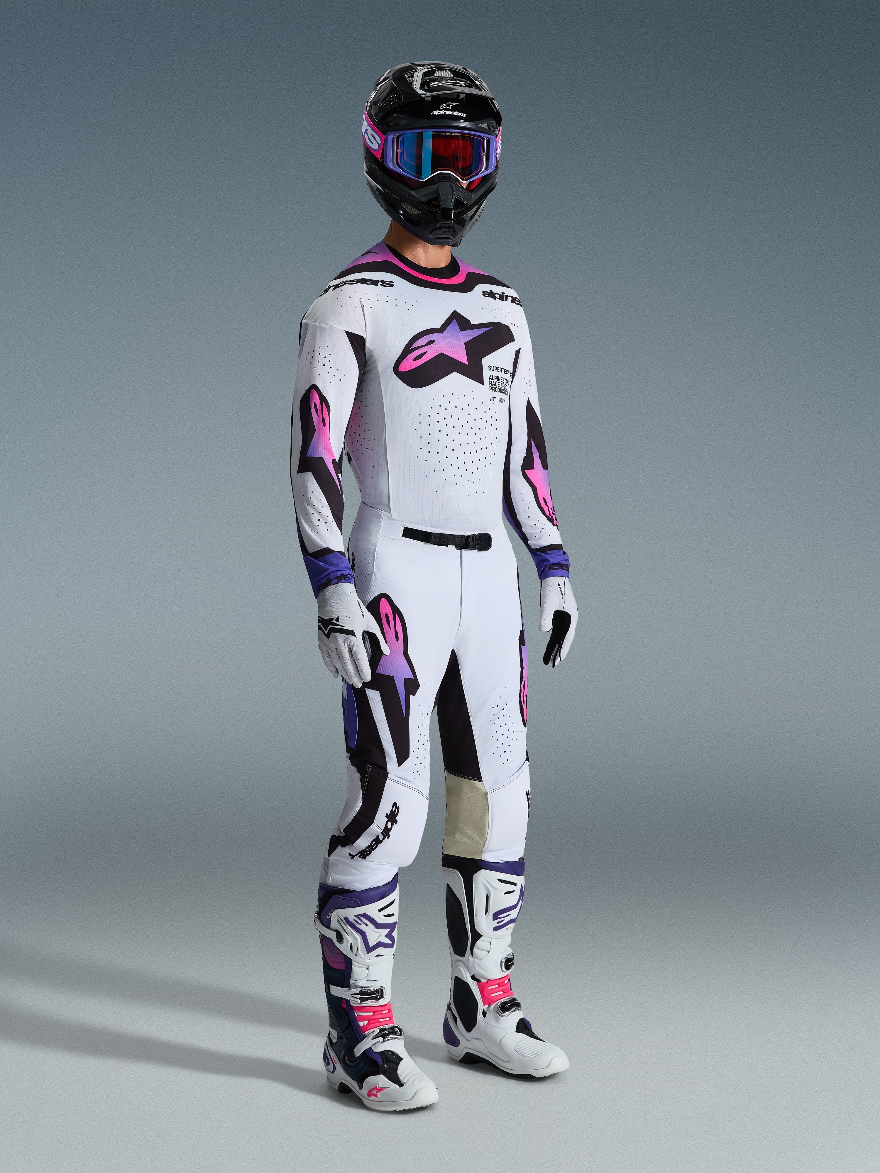 Motocross rider wearing complete racing gear, Alpinestars 2026 Supertech AFD Vista Gear Set, white jersey and pants with pink and purple gradient star logos, black accents, black helmet with purple frame goggles, white and black boots with pink details, coordinated professional motocross outfit