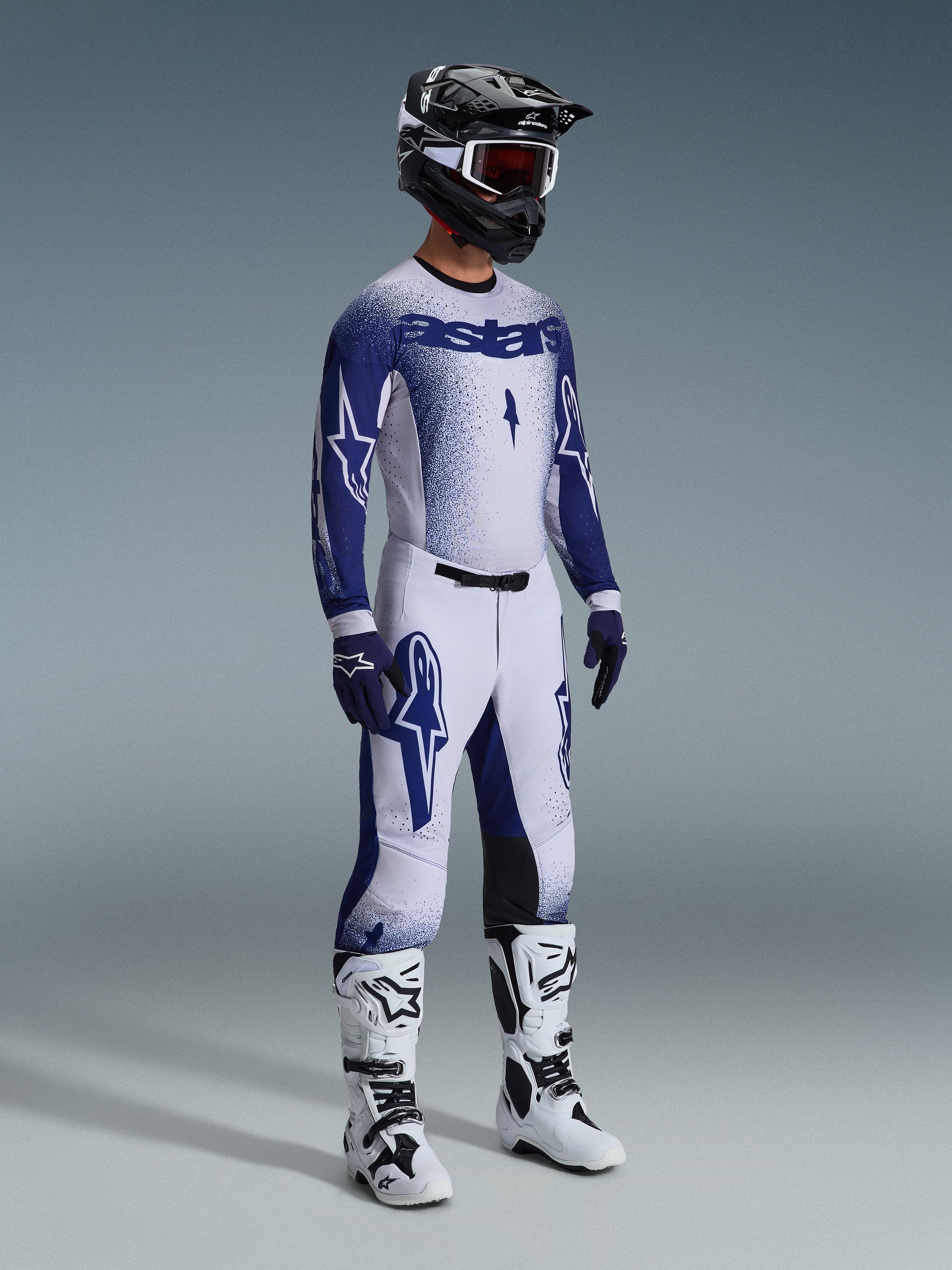 Motocross rider wearing 2026 Supertech Scenz Pants in Light Gray and Dark Blue, matching blue and white jersey with 'astars' branding and spray-paint effect, black helmet with white goggles, white and black motocross boots, coordinated professional off-road racing outfit.